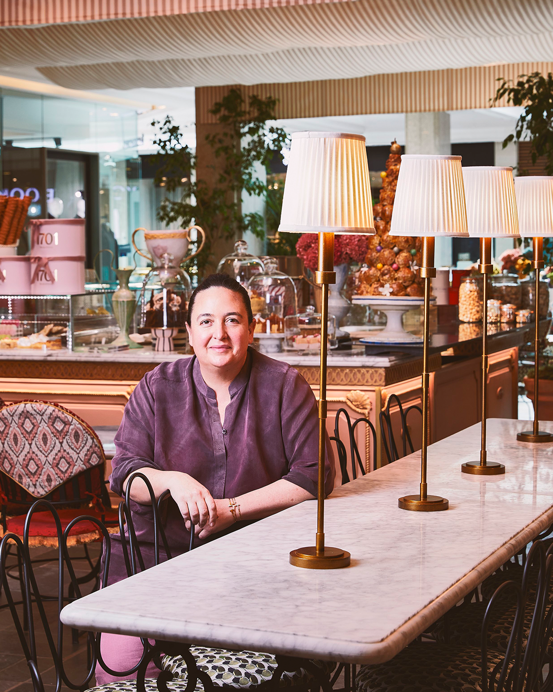 A woman sitting at a marble table in a decorated cafe or restaurant with a dessert display behind her and lamps on the table.