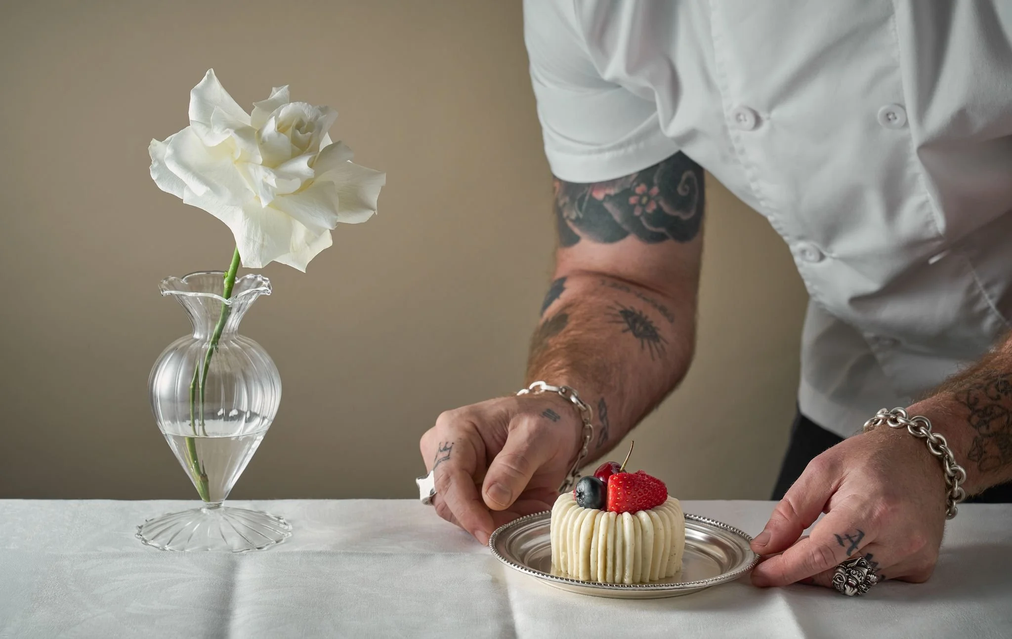 A person with tattoos and jewelry decorates a small cake with strawberries and blueberries. A white flower in a clear glass vase is on the table.