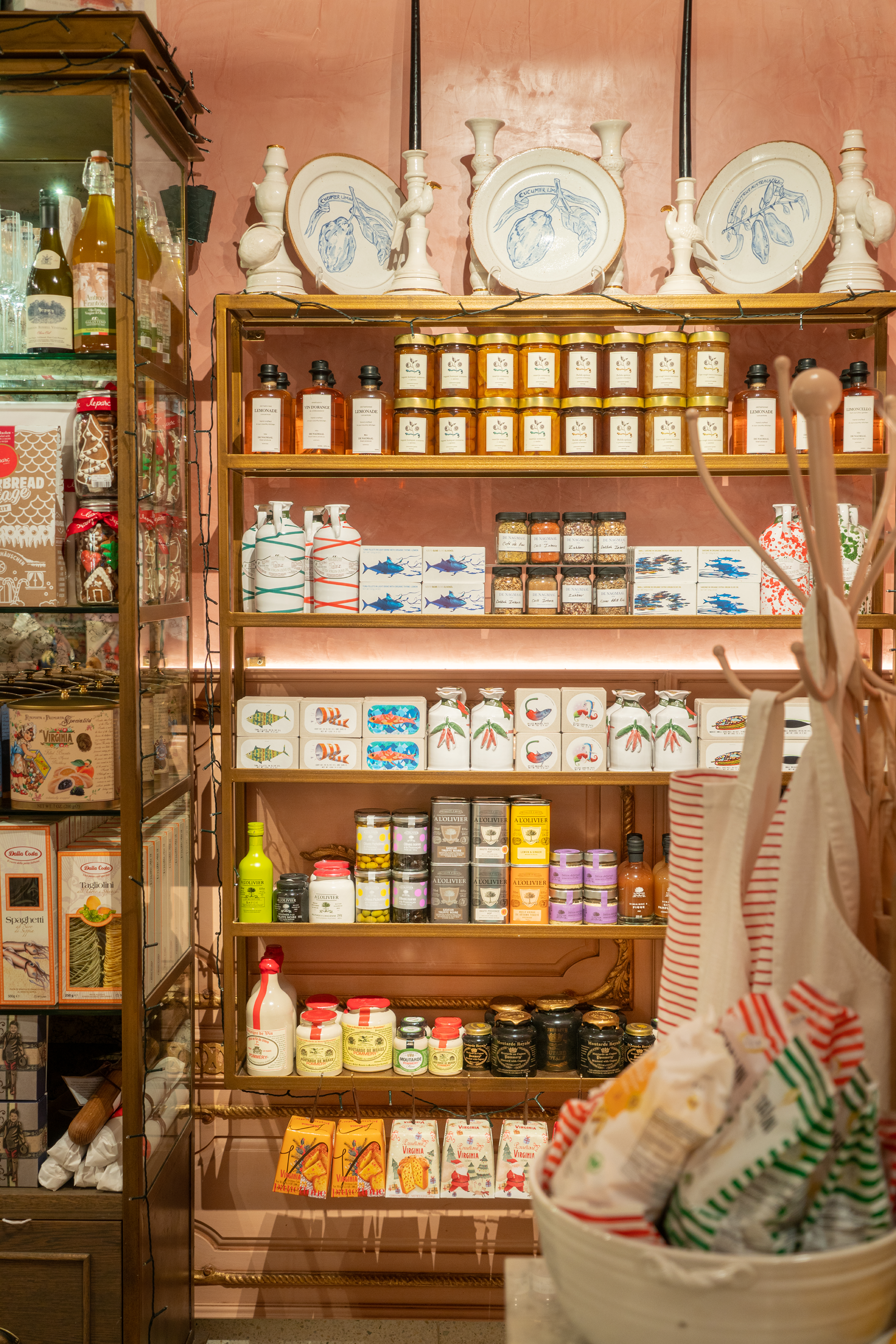 Display shelves with various jars and bottles of flavored spreads, sauces, and other gourmet food items, with decorative plates on the top shelf, set against a pink wall in a store.