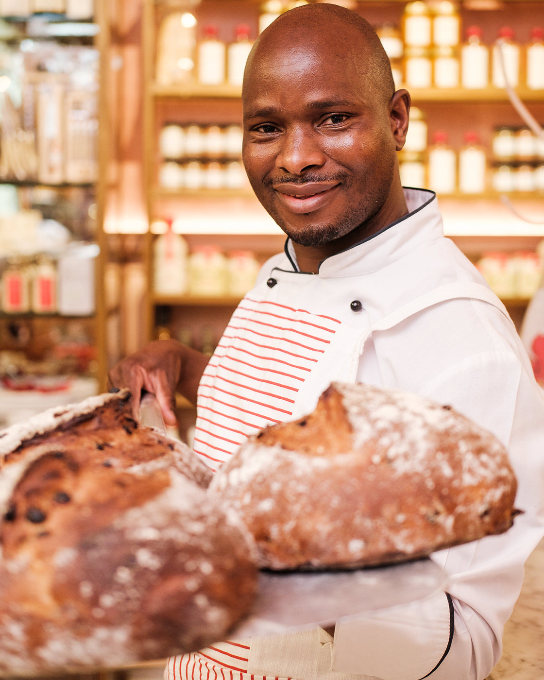 A smiling man in a white chef's uniform holding freshly baked bread in a bakery.