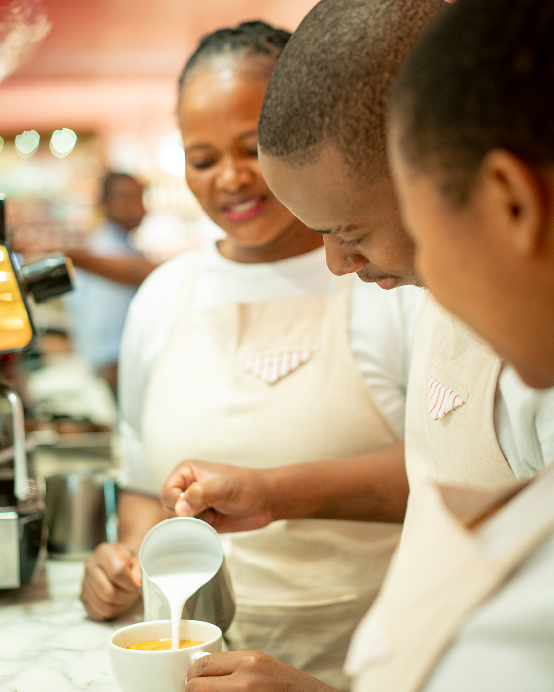 Three baristas in white uniforms and aprons preparing coffee behind a counter, with one pouring milk into a cup of coffee.