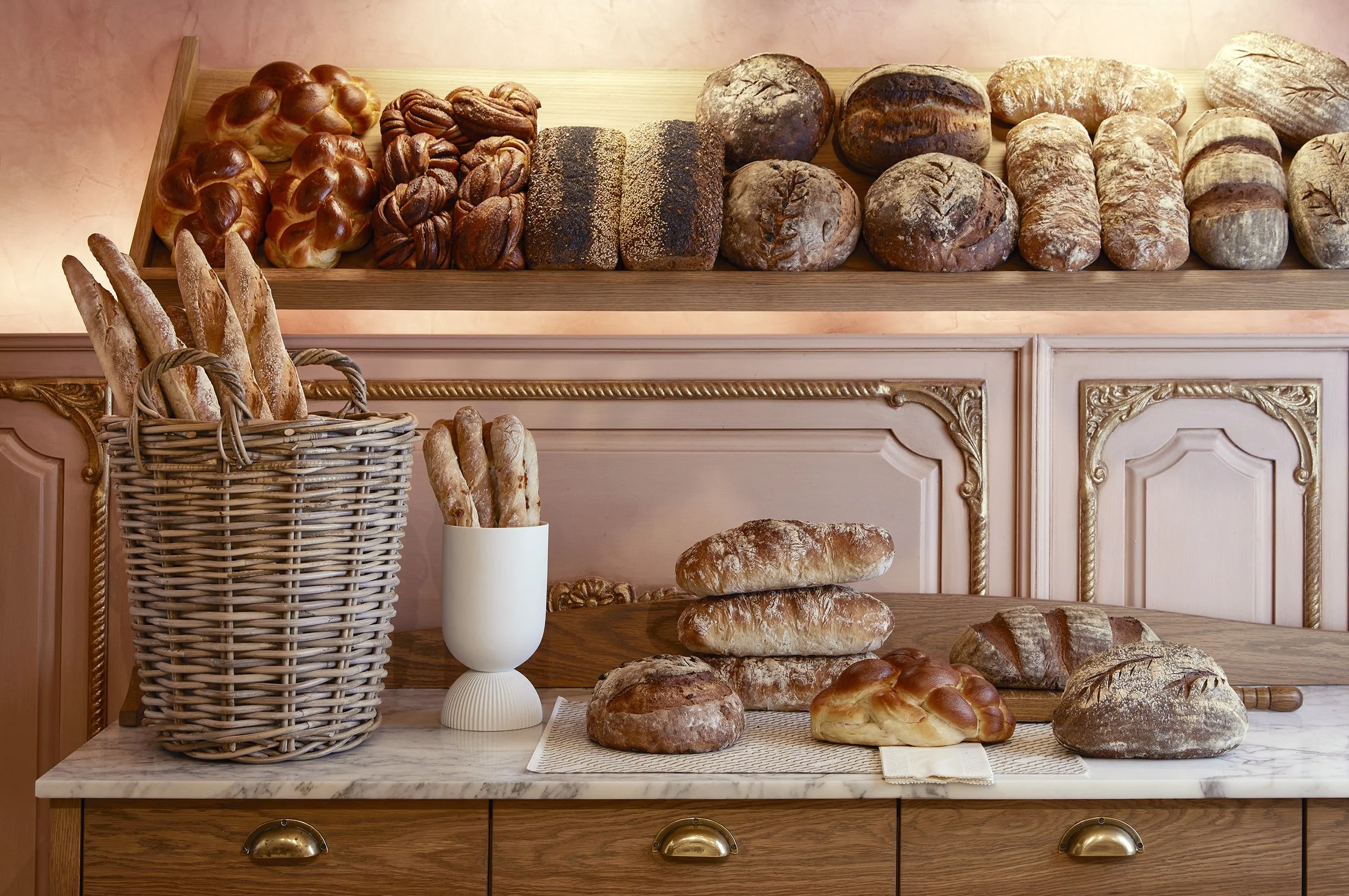 Display of various types of bread, including baguettes, rolls, and loaves, arranged on a wooden shelf and table in a bakery.