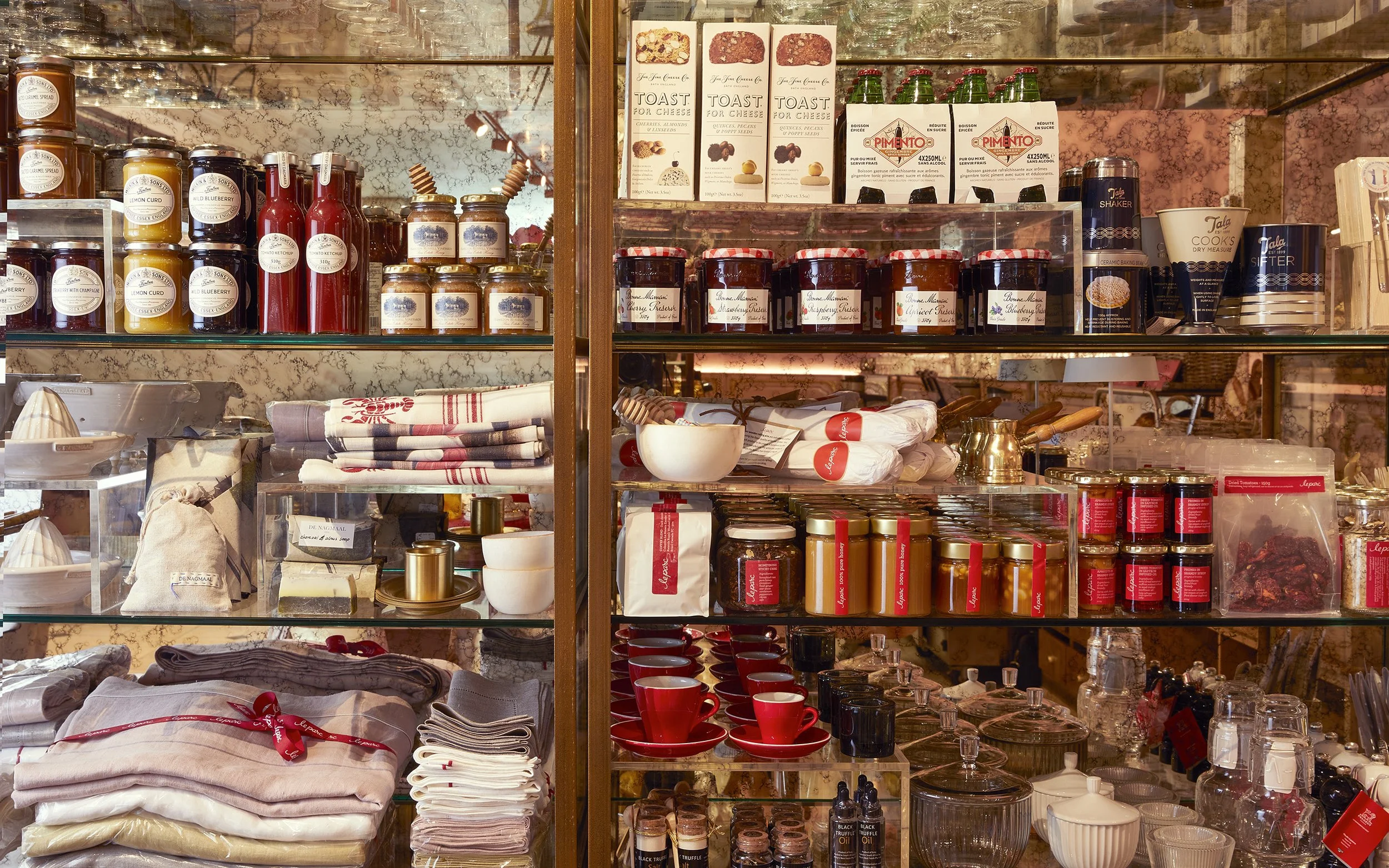 Display of various jars, bottles, and containers of food items, sauces, and kitchenware on glass shelves in a store. The background includes a marble wall and a variety of packaged products.