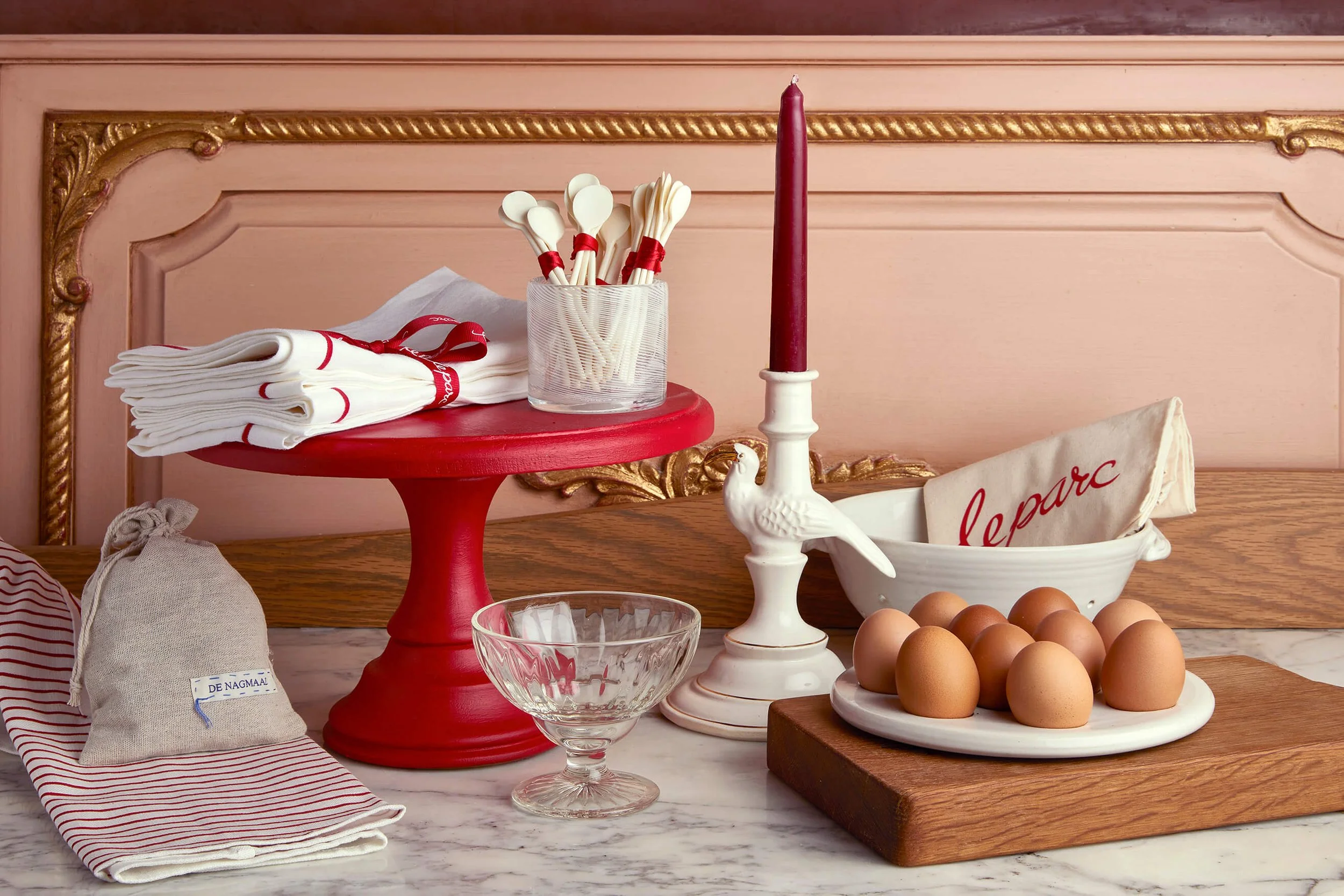 Kitchen scene with a pink wall, gold decorative trim, a red cake stand with white napkins, a glass container with white spatulas tied with red ribbons, a red candle in a white holder, a white swan-shaped dish with eggs, a clear glass bowl, and a white linen bag on a marble countertop.