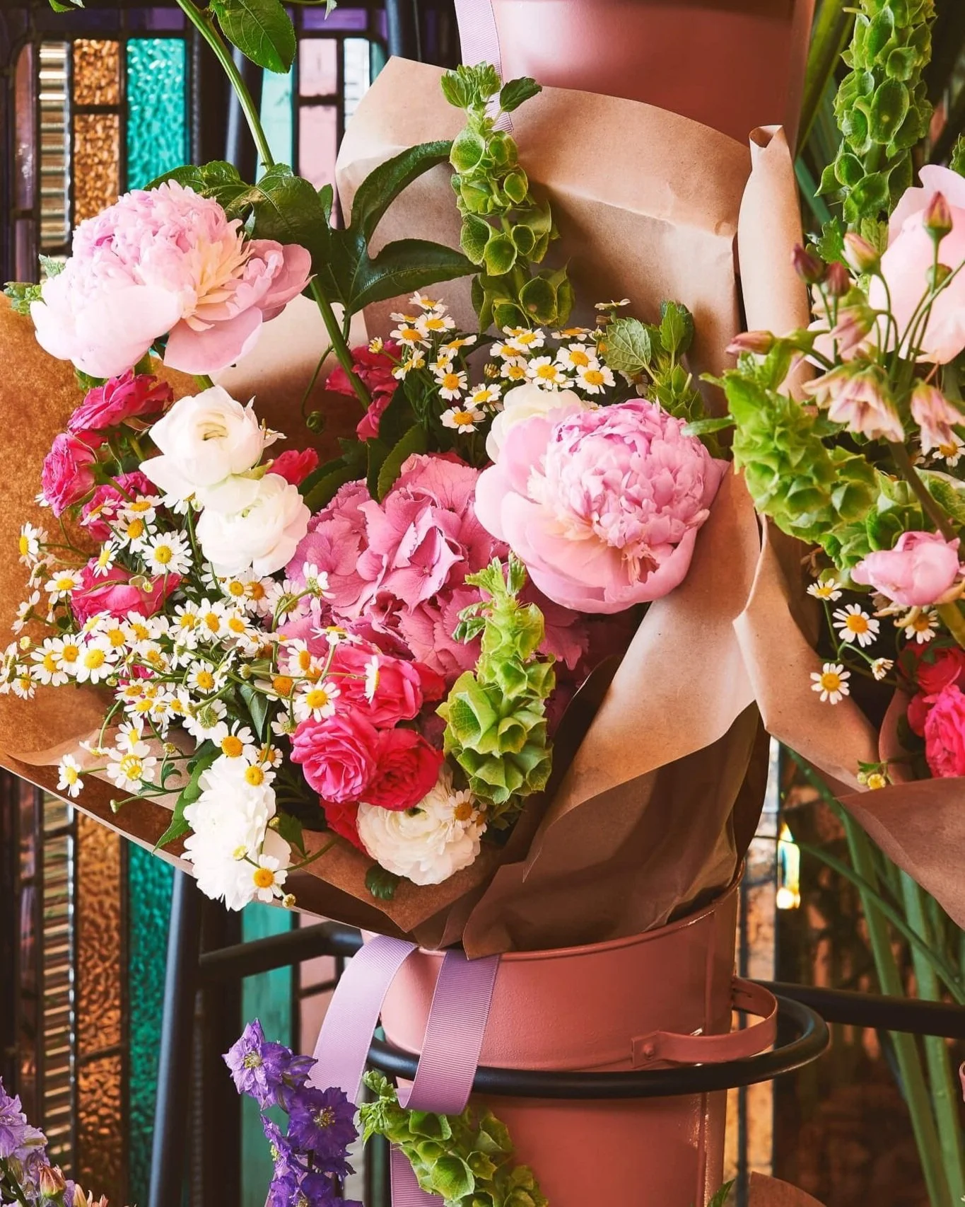 A wrapped bouquet of pink, white, and purple flowers with green foliage, tied with a pink ribbon, and set against stained glass windows.