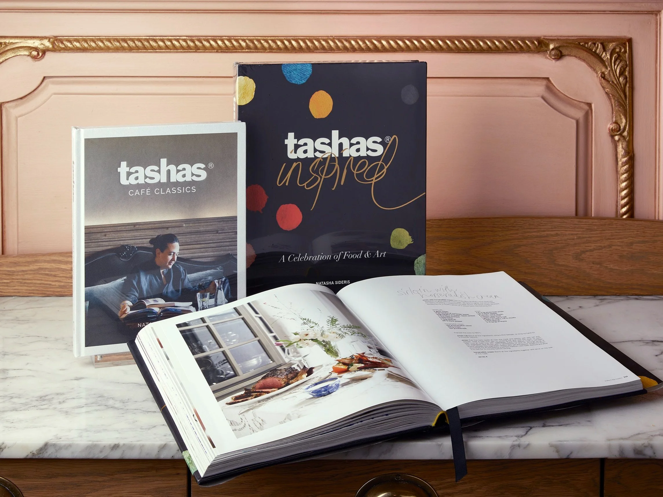 Open cookbook displaying food photographs and text on a marble countertop, with two promotional booklets standing upright behind it, on a wooden surface with ornate pink and gold decor in the background.