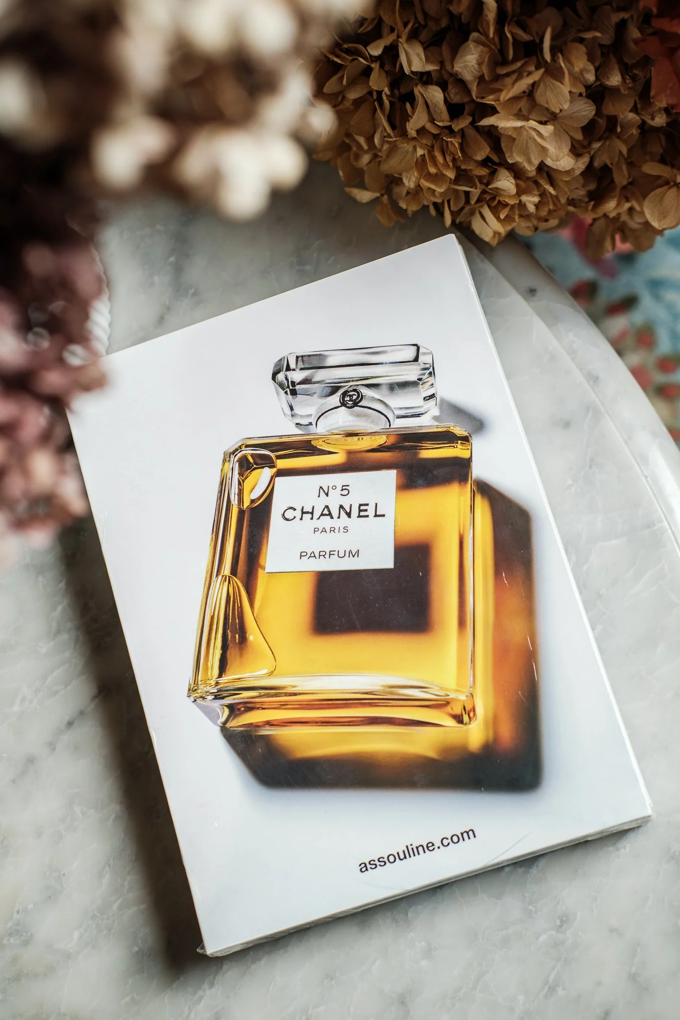 A box of Chanel No. 5 perfume on a marble surface, with dried hydrangea flowers in the background.