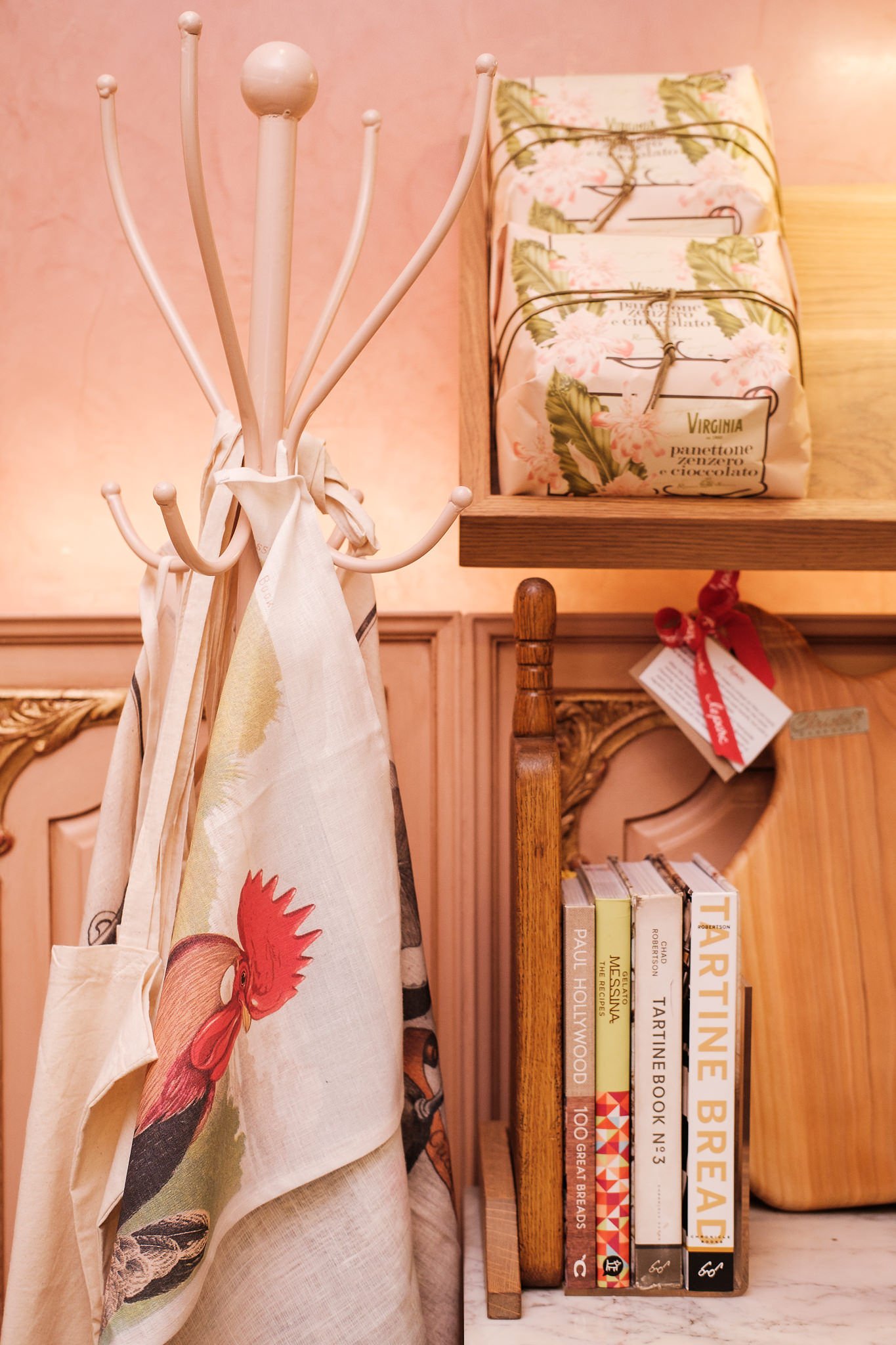 A white coat rack with a scarf hanging, next to a wooden bookshelf holding several books. The background has a pink wall.