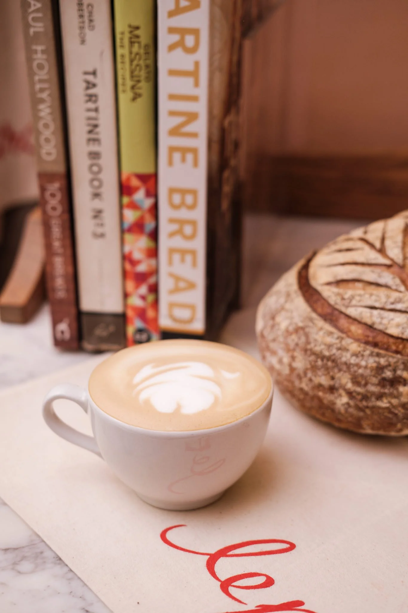 A white ceramic coffee cup filled with latte art on a white surface with a background of books and a decorative object.