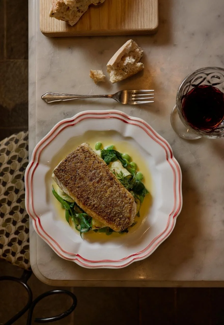 A plate with a piece of cooked fish garnished with green herbs, served on a bed of green vegetables with a yellow sauce. To the right, there is a glass of red wine. Above the plate, a piece of bread, a fork, and a small cutting board with more bread are visible.