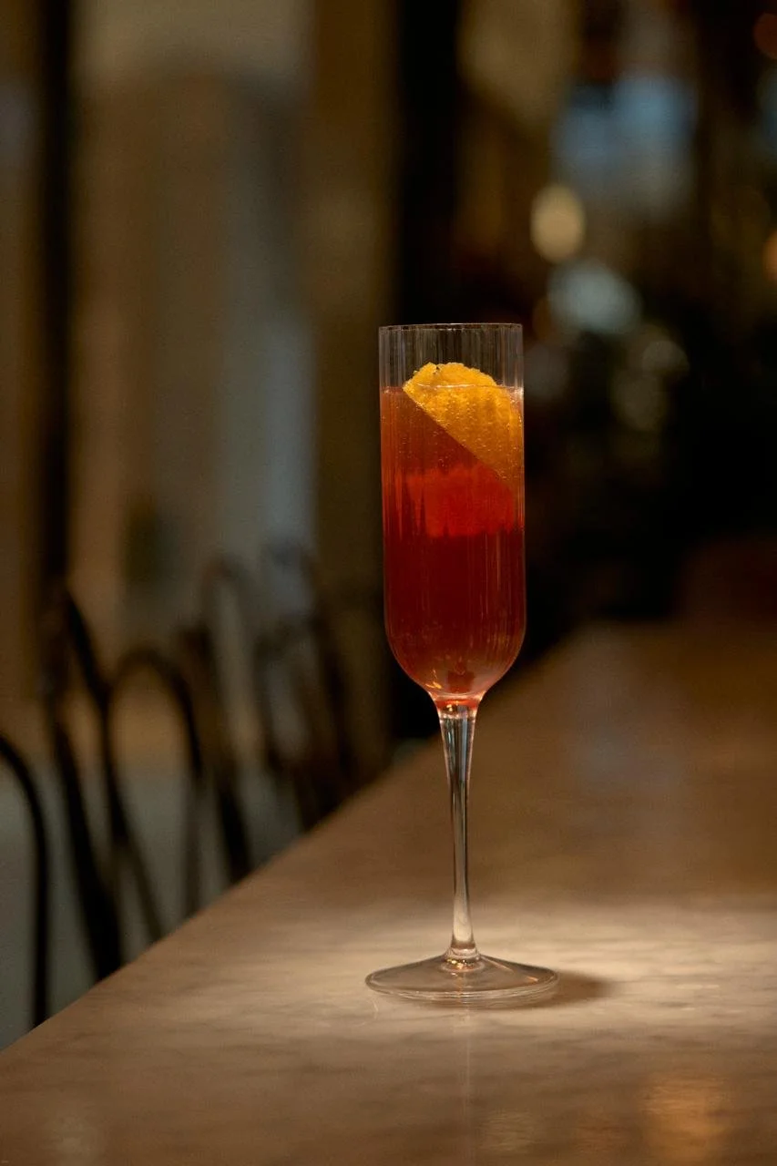 A tall champagne flute filled with red-colored beverage, garnished with a twist of orange peel, on a wooden bar counter with a blurred background.