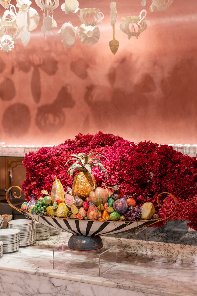 Decorative Fruit Display with a variety of painted, colorful fruits including a gold pineapple, pears, grapes, and other fruit, set in a large ornate bowl on a marble surface, with a backdrop of red flowers and a pinkish wall with hanging paper decorations.