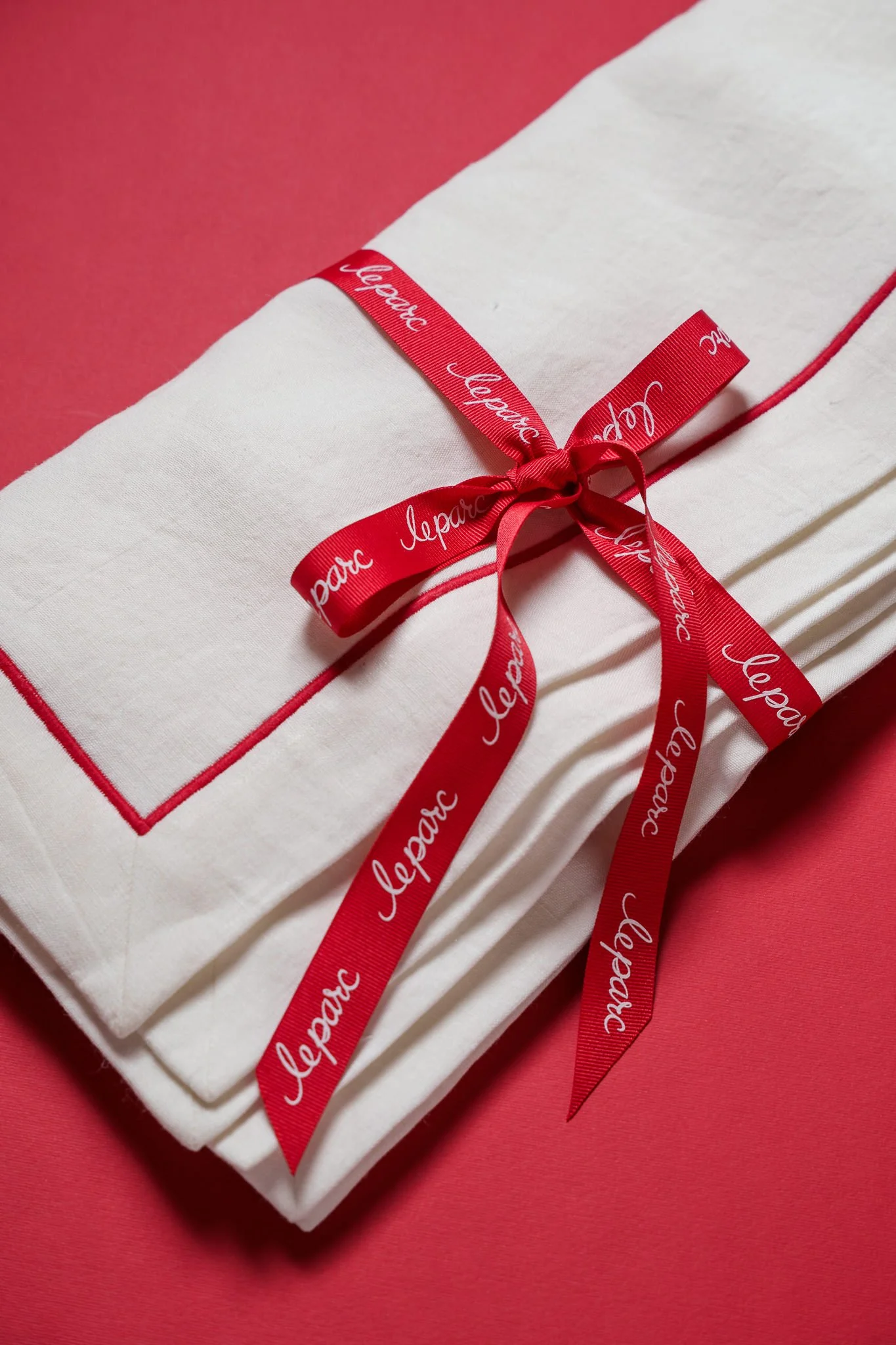 A neatly folded white napkin with a red ribbon tied in a bow, with the word 'Leparc' printed repeatedly in white on the ribbon, placed on a pink background.