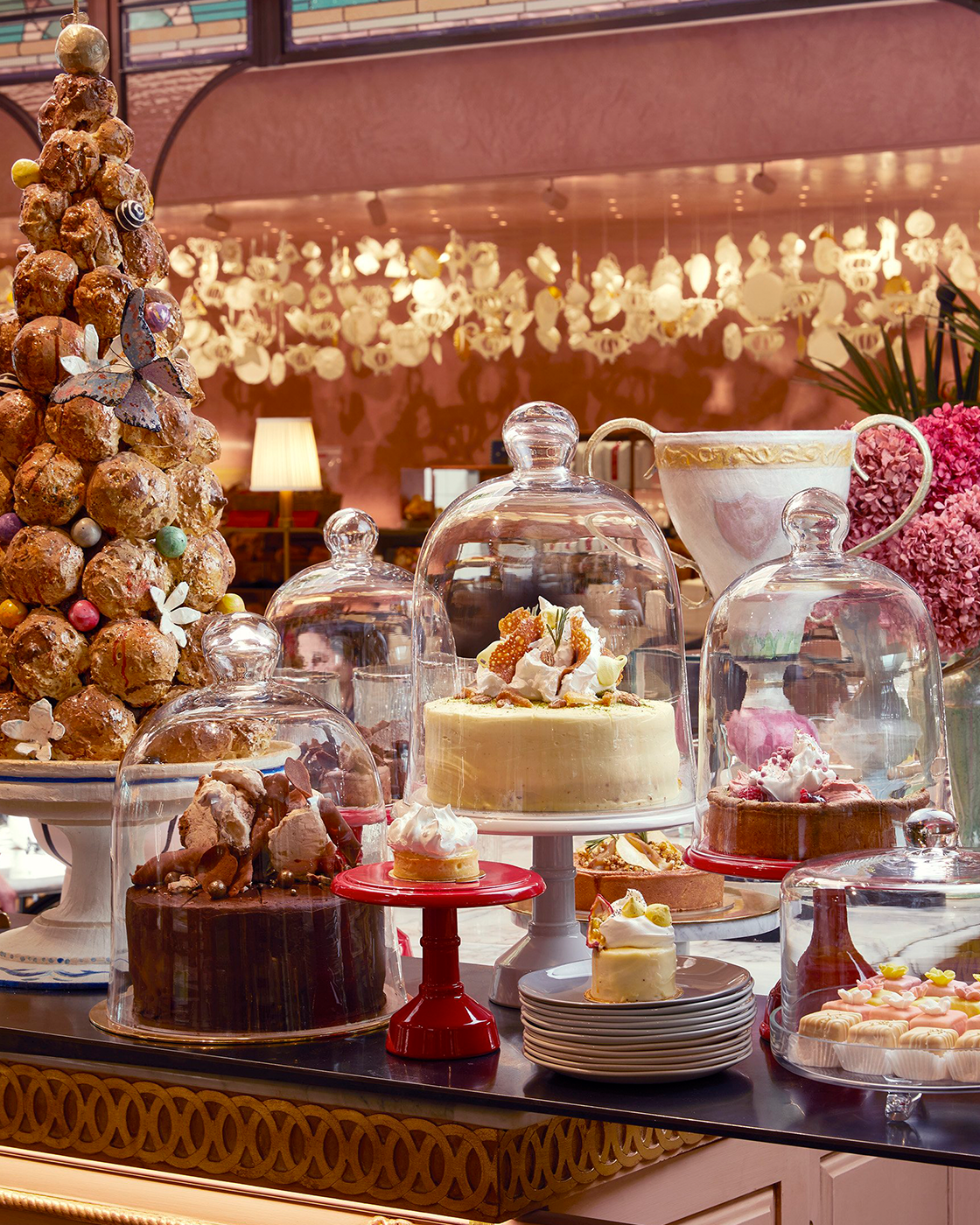 Display of various cakes and desserts inside glass domes at a bakery, with a Christmas tree decorated with chocolates and ornaments to the left.