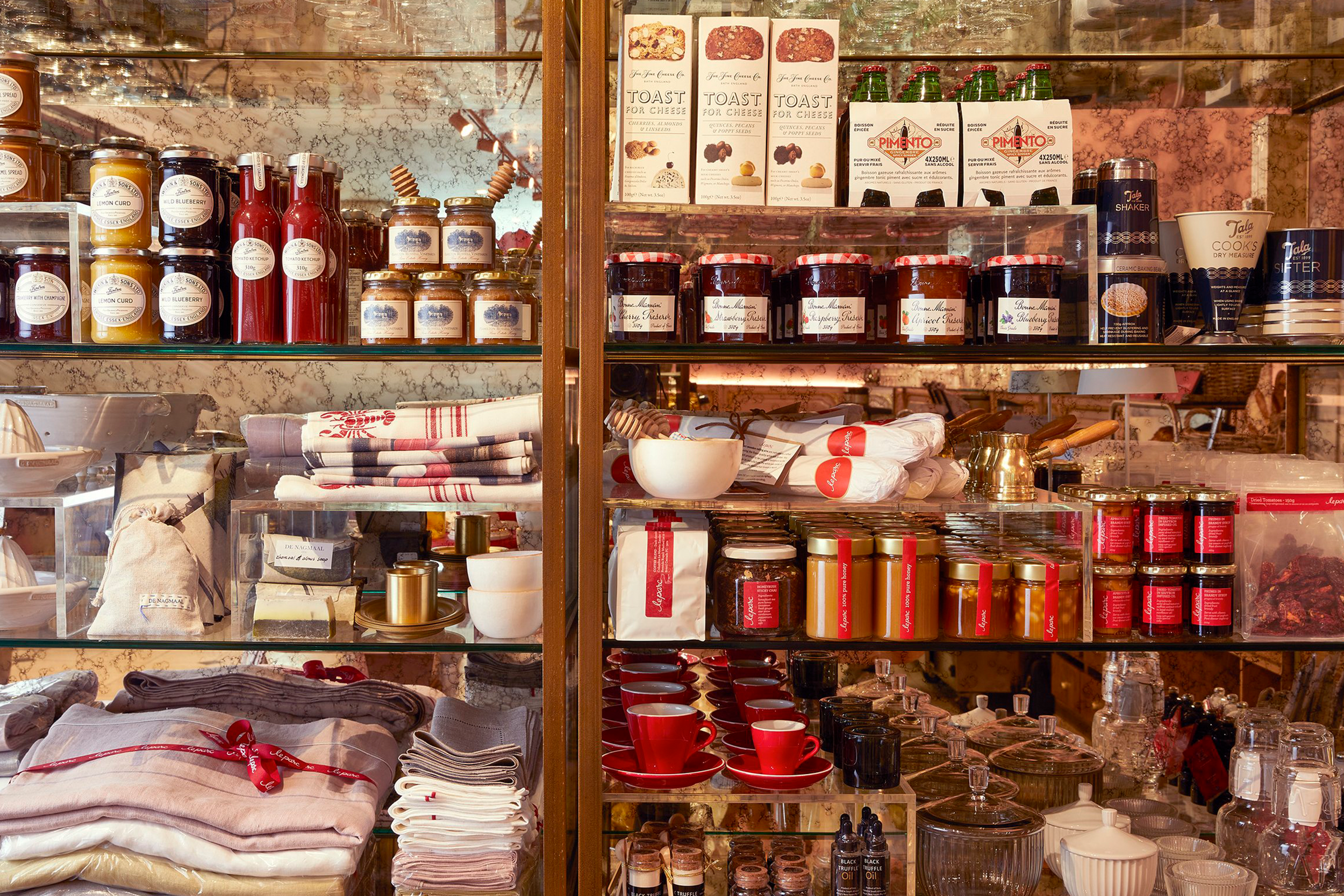 Display shelf with various jars of jam, bottles of syrup, boxed cookies, cups, and kitchen towels.