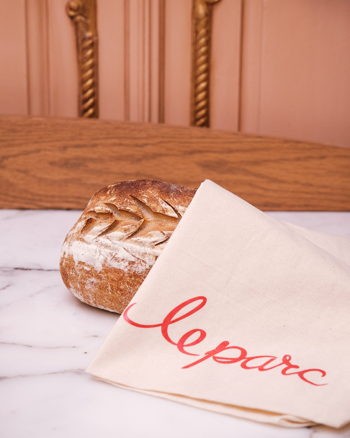 A loaf of bread with a decorative carved top, placed on a marble surface next to a white napkin with red cursive writing that says 'le pain'.