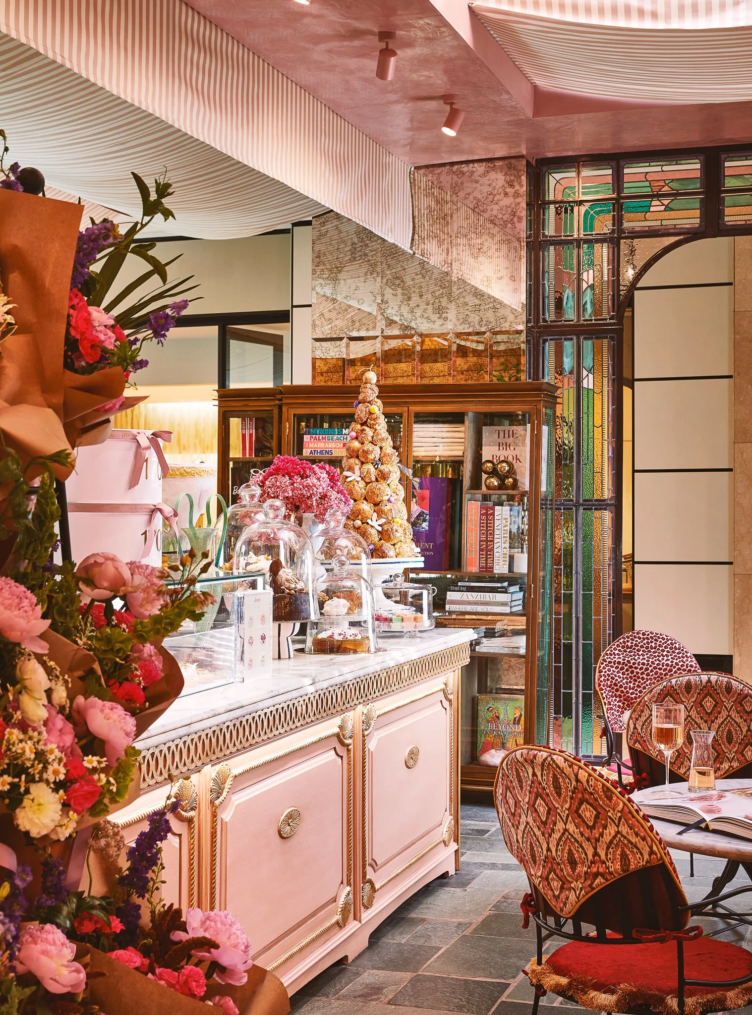 Interior of a café or restaurant with pink furniture, floral arrangements, a decorated pastry display, and stained glass windows.