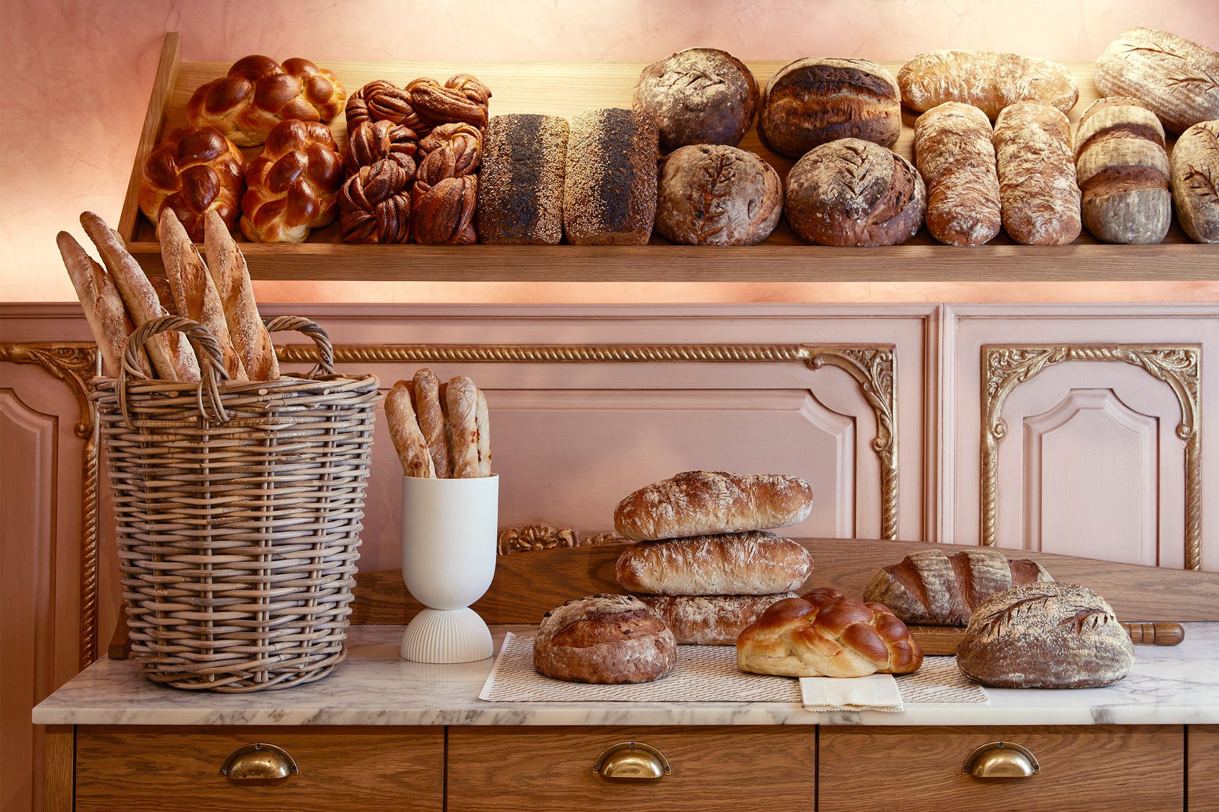 Display of various breads on a wooden and marble table, with a woven basket of baguettes and a white vase of breadsticks, set against a pink ornate wall.
