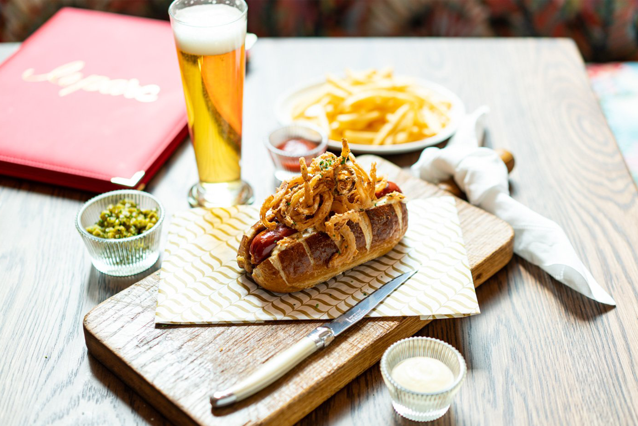 Hot dog with fried onions on a bun, surrounded by small bowls of chopped green peppers, white sauce, and ketchup, set on a wooden cutting board on a table with fries and a glass of beer in the background.