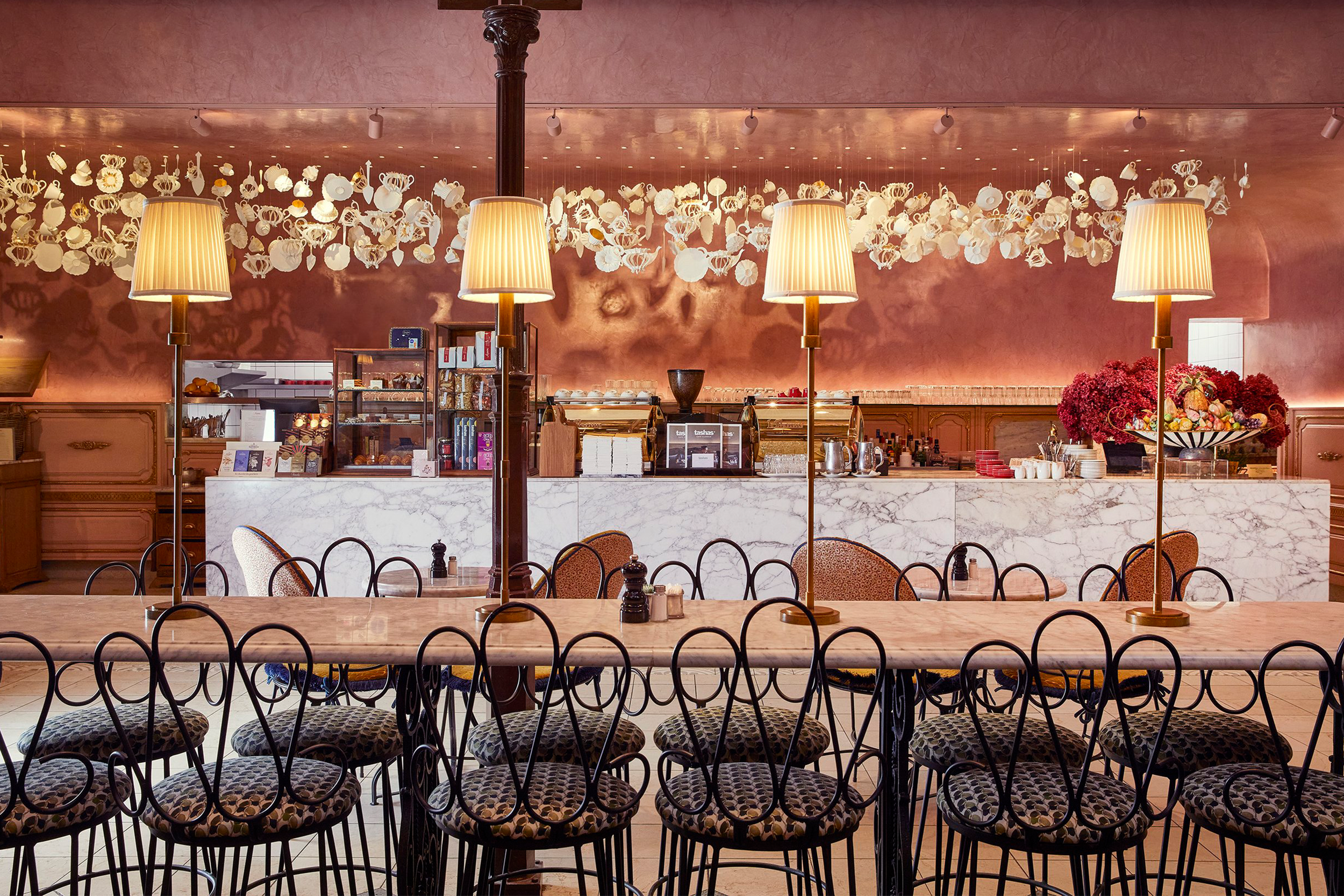 Interior of a restaurant or cafe with a marble-topped counter, three table lamps, and a pink wall decorated with hanging white paper flowers and cups. There are chairs with patterned cushions around the tables in foreground.