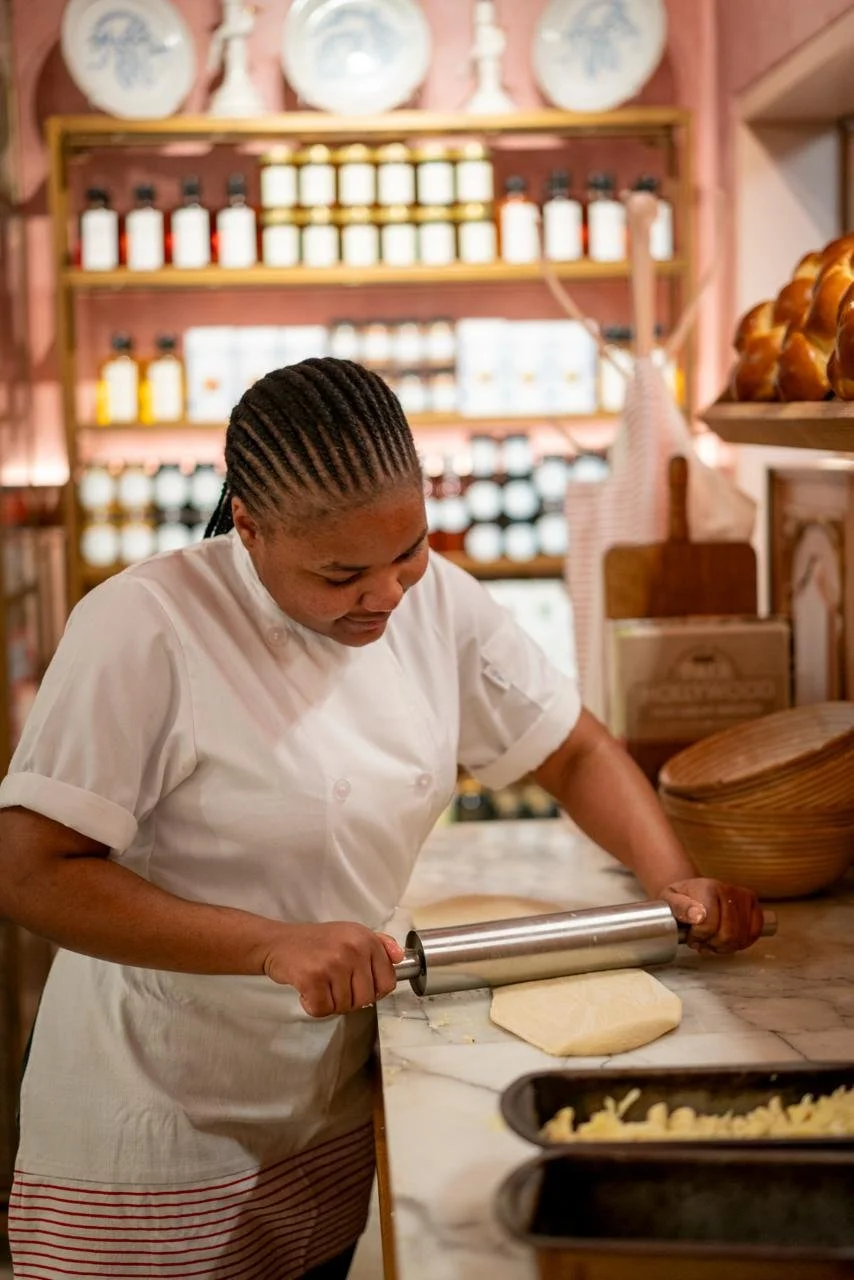 A woman in a white chef's coat rolling out dough with a rolling pin on a marble countertop in a bakery or kitchen.