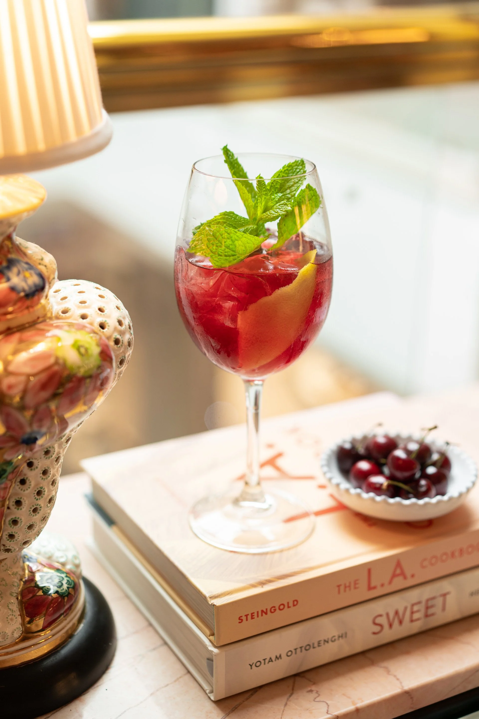 A glass of red iced beverage garnished with mint leaves and lemon slices, placed on a white box of books, with a small bowl of cherries nearby.