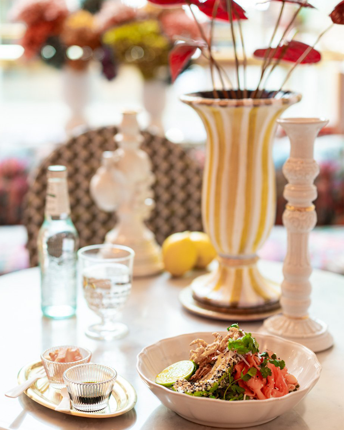 A table with a colorful salad, a lime wedge, lemon, and condiments, set among decorative vases and candlesticks, with a blurred floral background.