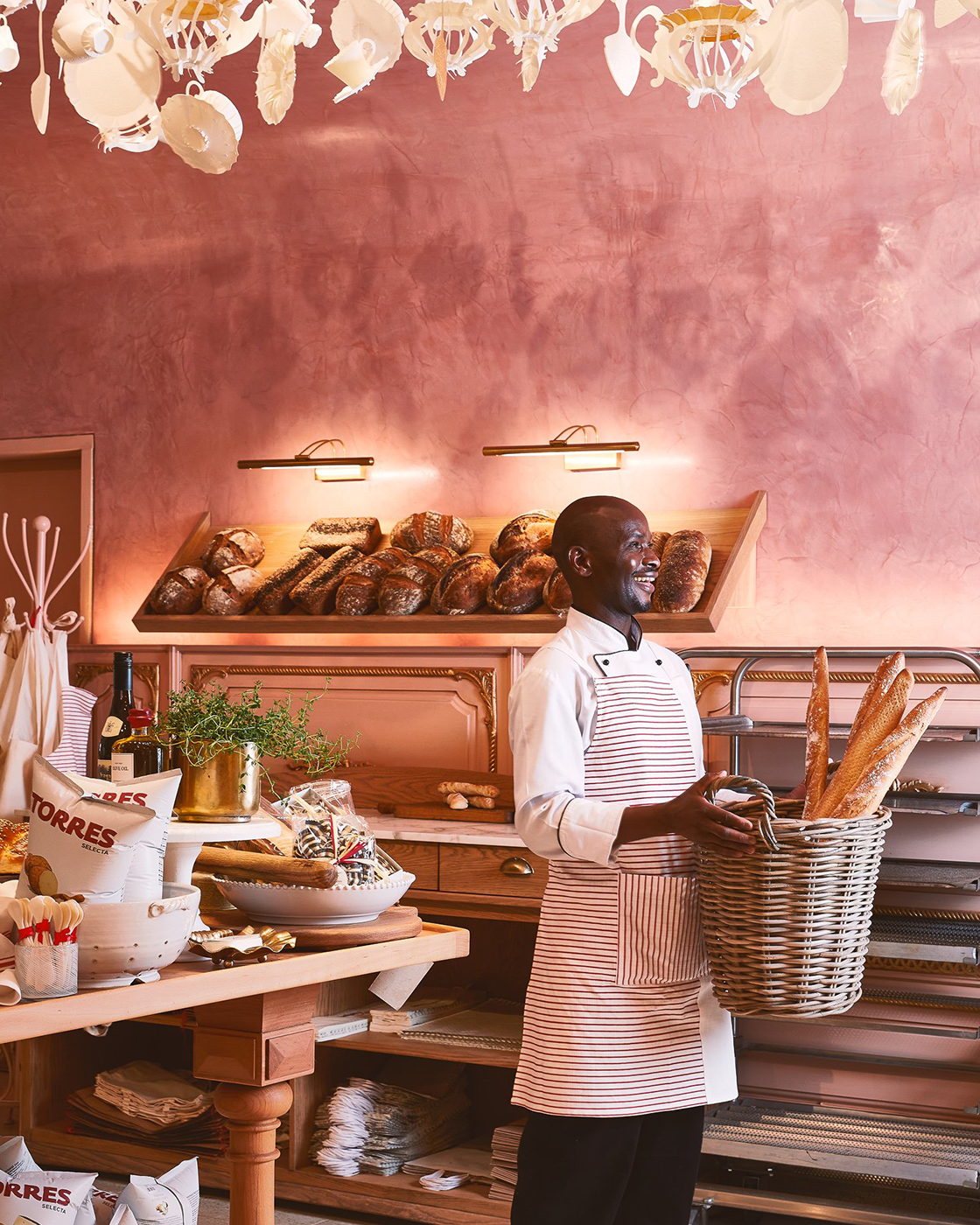 A smiling baker standing in a bakery, holding a basket of baguettes, with racks of bread and baked goods, pink textured wall, and decorative lighting in the background.