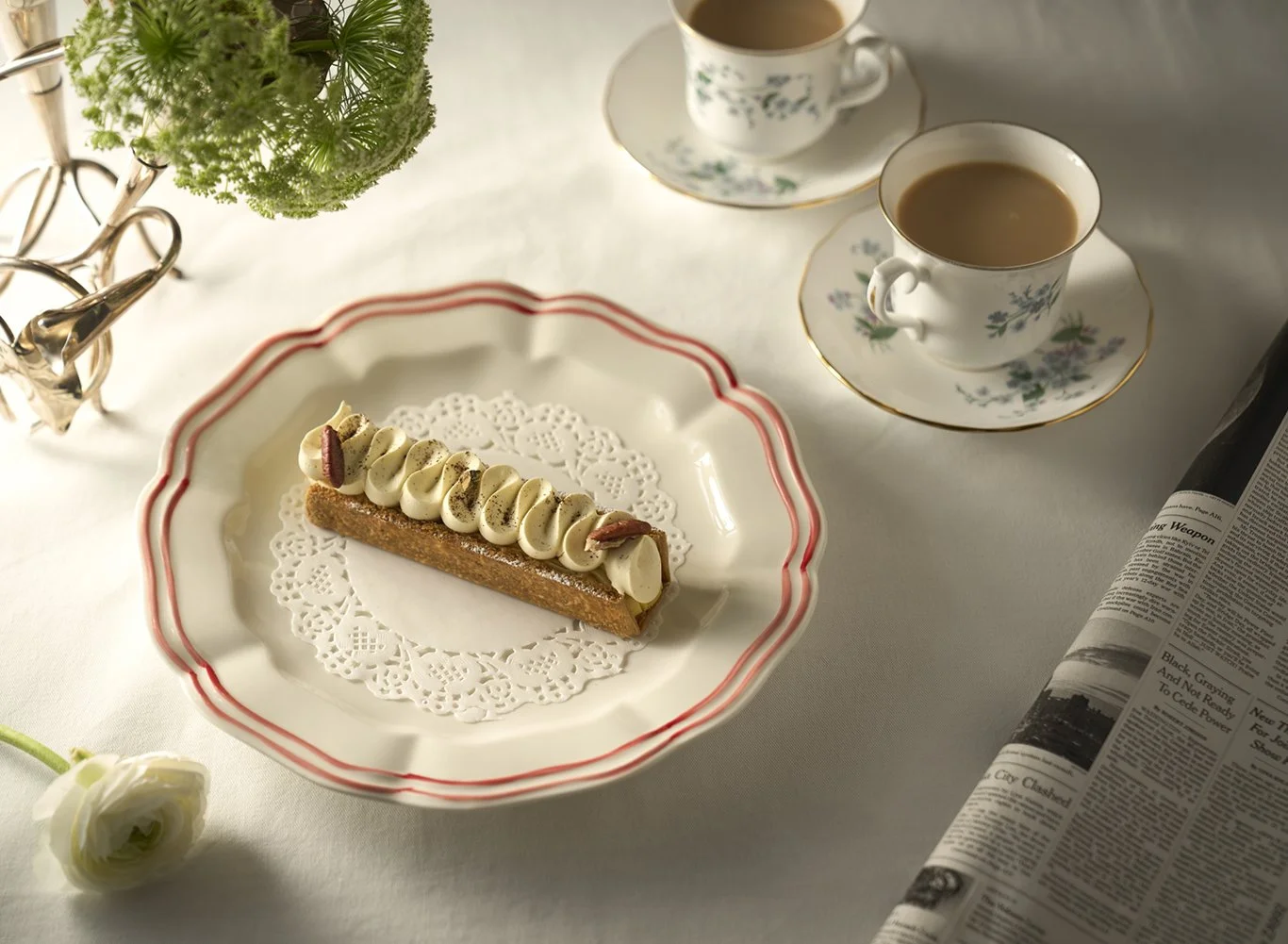 A table with a white tablecloth featuring a dessert, two cups of tea, a newspaper, a flower, and a plant. The dessert is a rectangular tart with white frosting and small decorations on top, placed on a decorative plate with a doily underneath.