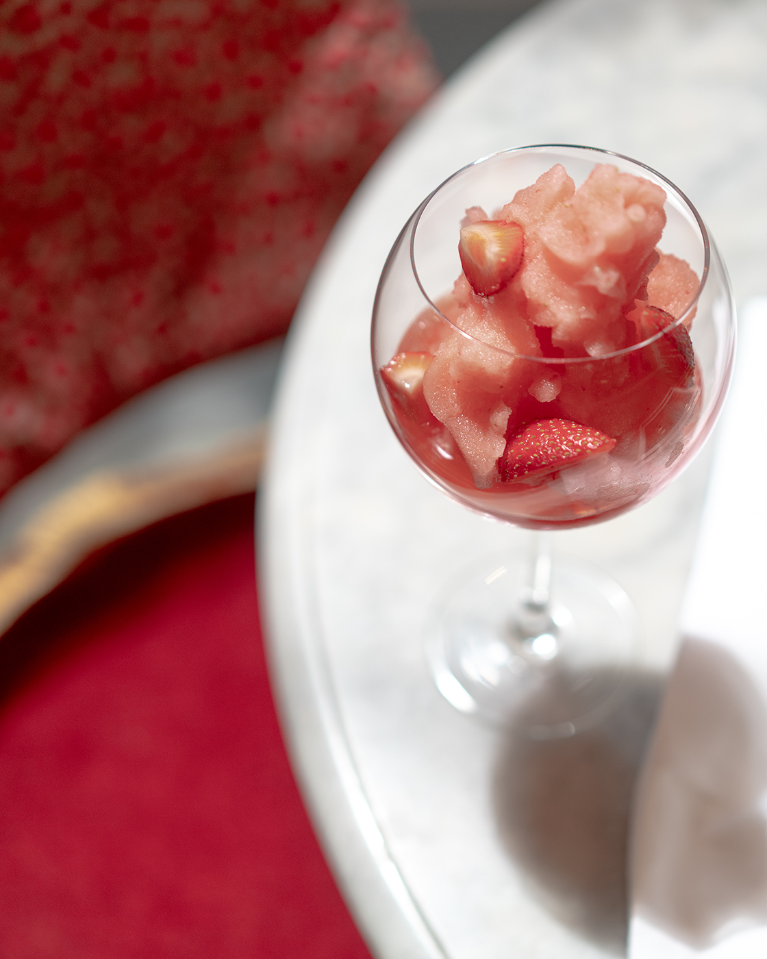 A glass of pink strawberry sorbet with fresh strawberry pieces on top, placed on a white round table with a blurred watermelon in the background.