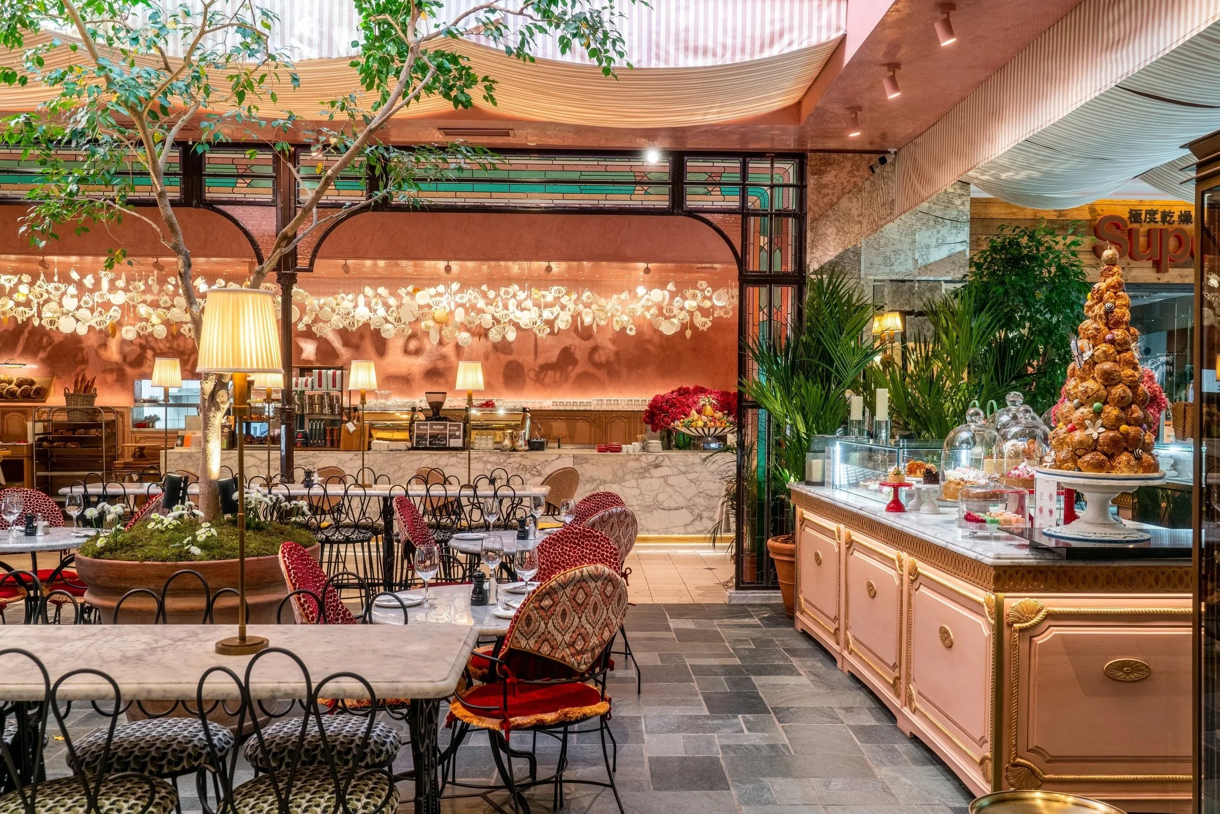 Interior of a cafe or restaurant with tables, chairs, a decorative tree, plants, and a display of desserts and pastries. The decor features warm lighting, pink accents, and artistic wall decorations.