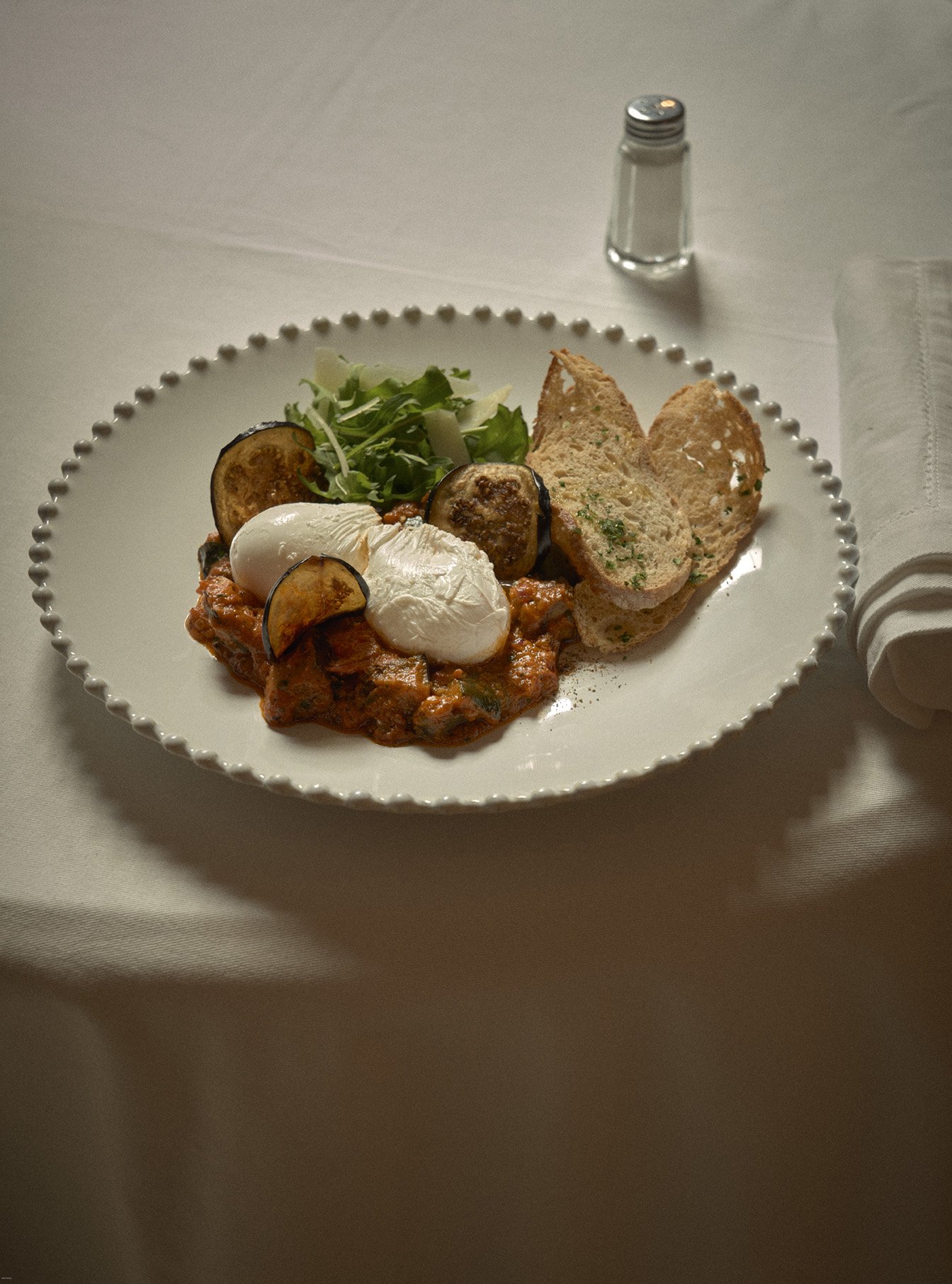 Plate of eggs benedict with poached eggs, roasted eggplant slices, mixed greens, and slices of toasted bread, with a salt shaker in the background.