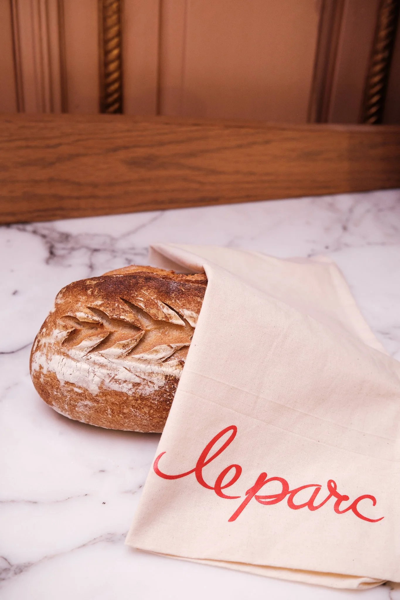 A loaf of crusty bread partially wrapped in a white napkin, resting on a white marble surface with a wooden background.