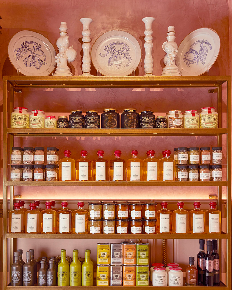 Shelf displaying jars of preserves, bottles of olive oil, and ceramic platters with botanical illustrations on a pink wall.