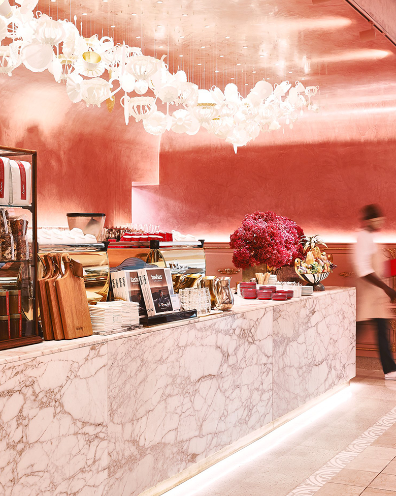 A polished marble counter with kitchen items, books, and a large pink flower arrangement against a pink metallic wall, with a decorative white chandelier hanging above and a person walking by.
