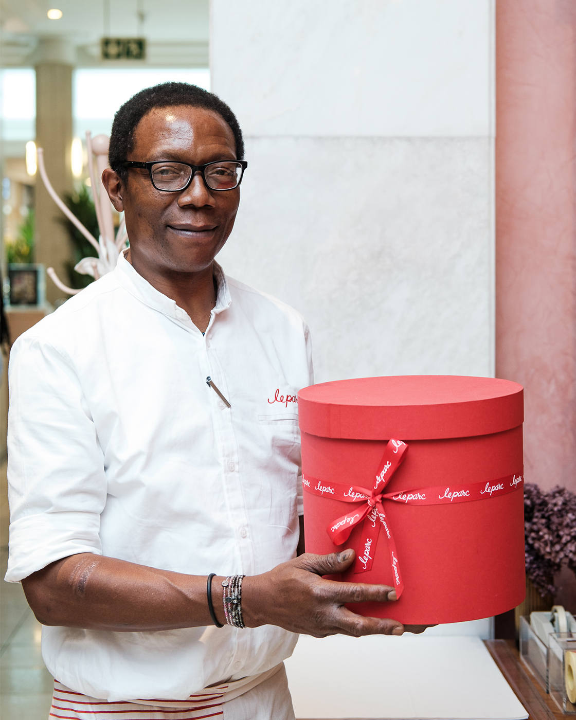 A man with glasses and a white shirt holding a large red gift box with a ribbon that says 'lepore' in an indoor setting.