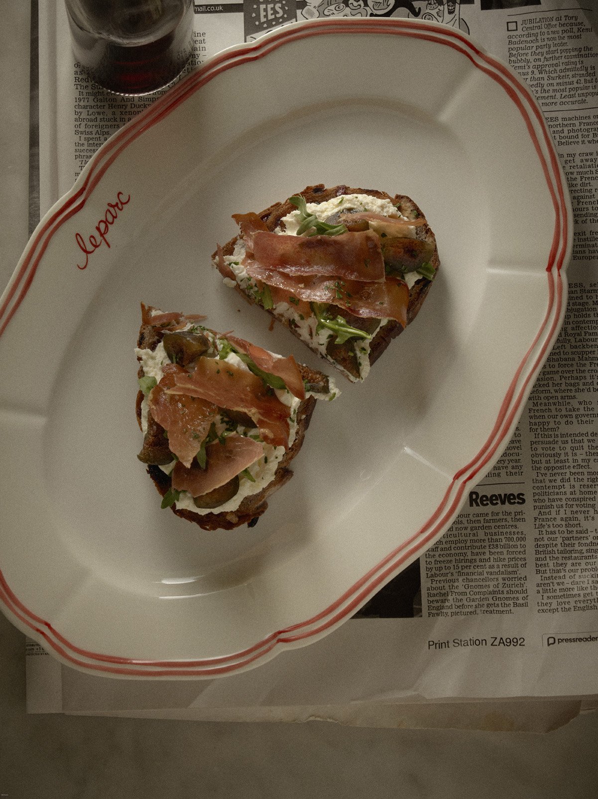 Two open-faced sandwiches with prosciutto, arugula, and cheese on toasted bread, served on a white oval dish with red border, on newspaper print surface, with a glass of dark beverage in the background.