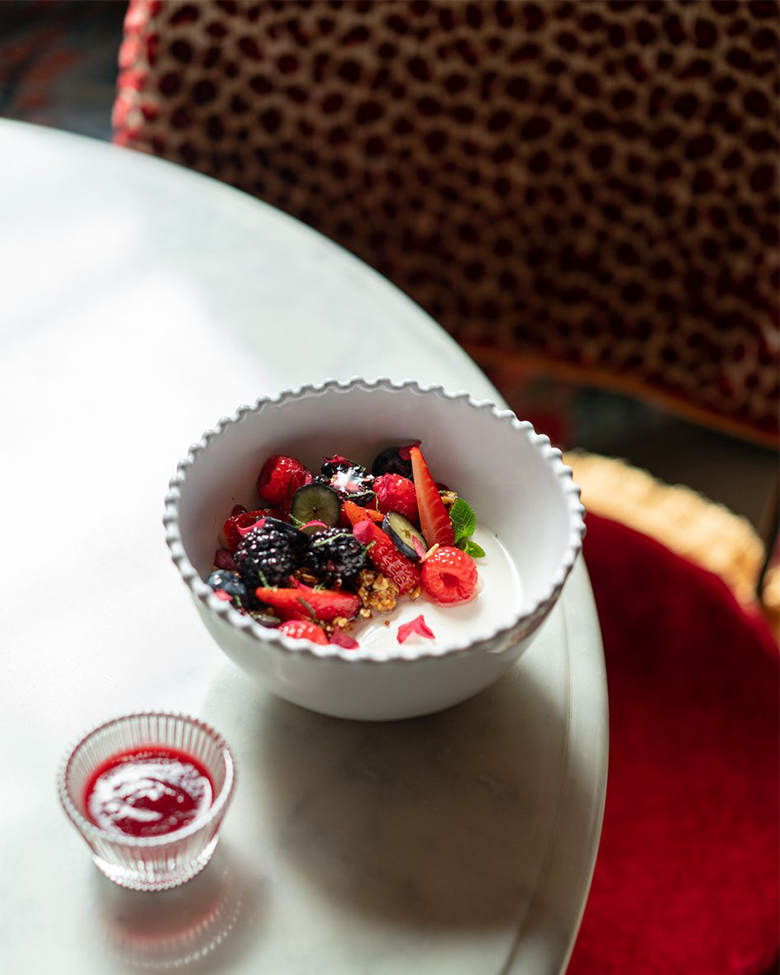 A white bowl of mixed berries placed on a white marble table, with a small glass container of berry syrup beside it. Part of a red and patterned chair and a red carpet are visible in the background.