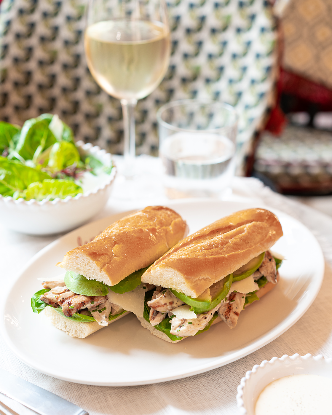 Two chicken salad sandwiches on a white plate with lettuce, green pepper, and cheese, with a side salad, a glass of white wine, and a glass of water in the background.