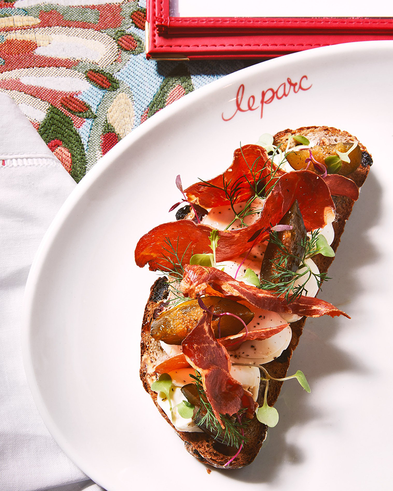 Open-faced sandwich with smoked salmon, cured meat, microgreens, and a poached egg on toasted bread on a white plate with red cursive writing.