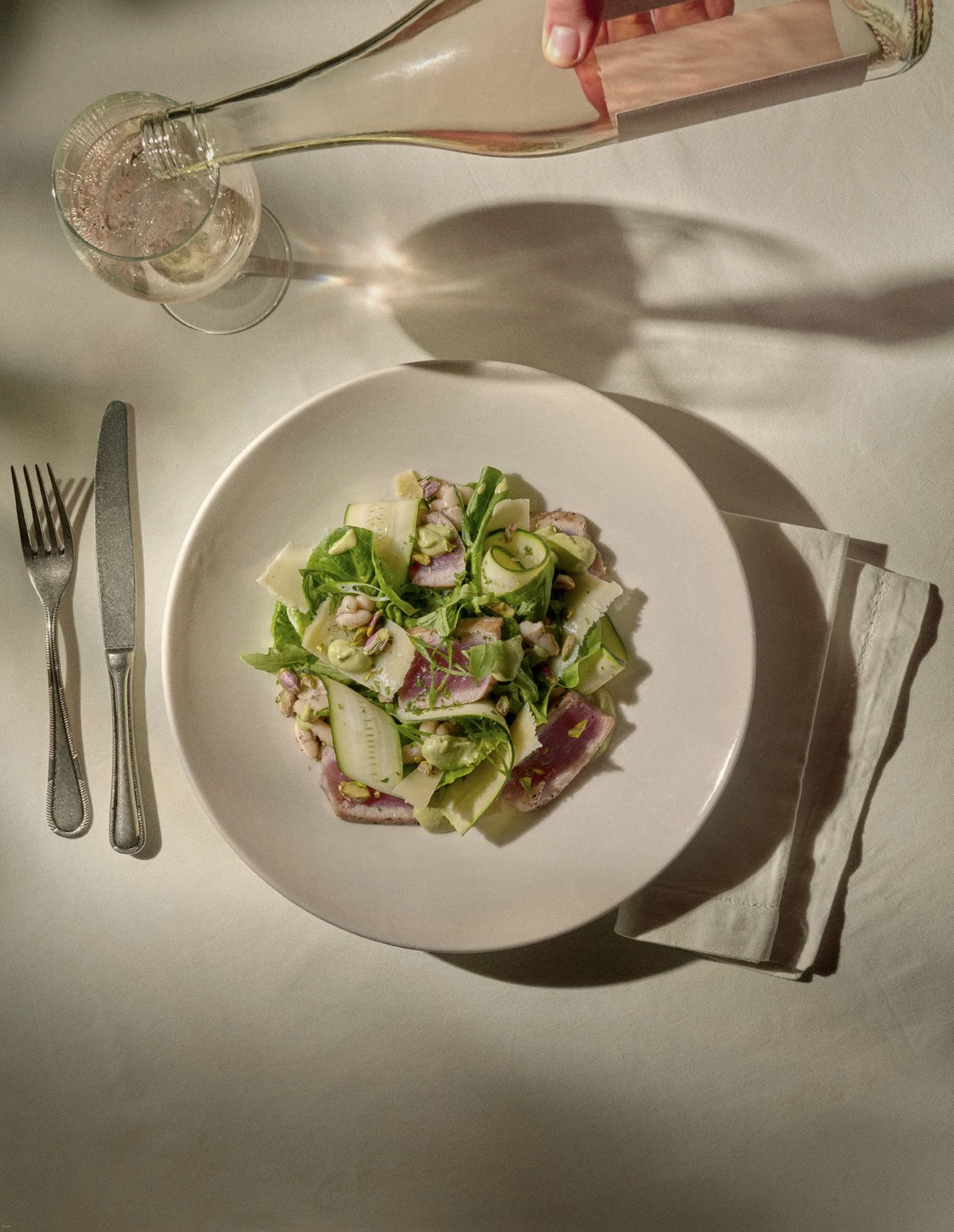 A plate of salad with leafy greens, sliced cucumbers, shaved cheese, and pink sliced meat. There is a glass of sparkling water and a wine bottle being poured into the glass. Silverware is placed on either side of the plate, on a white tablecloth with a napkin to the right.