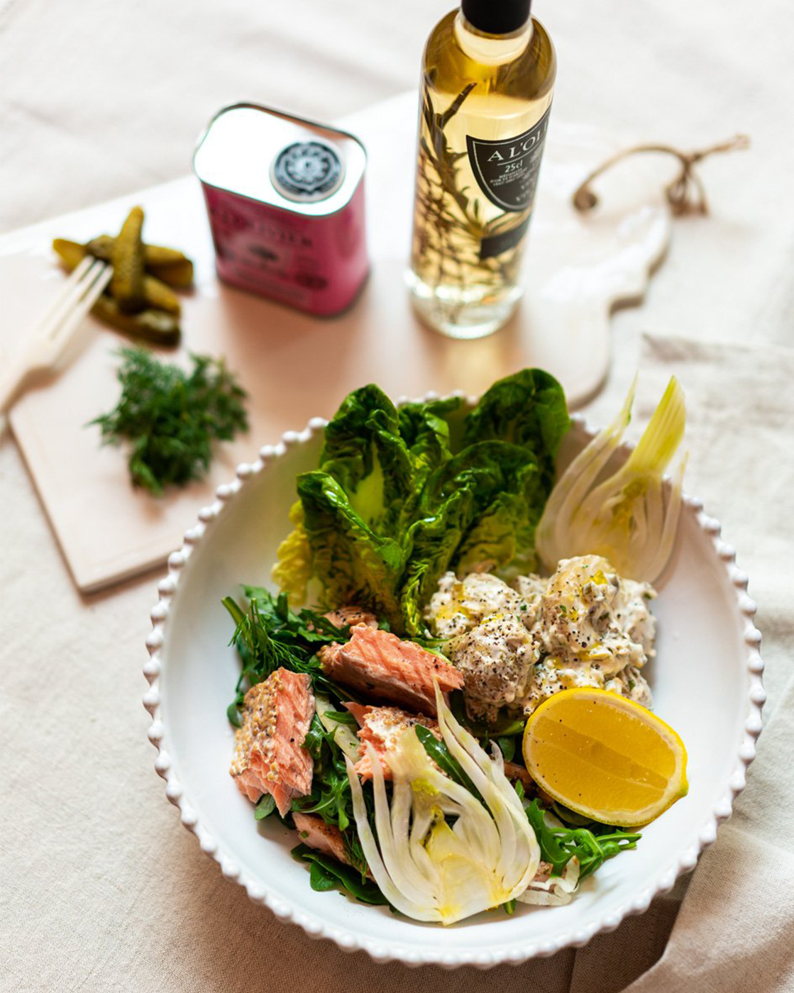 Salad with lettuce, salmon, lemon wedge, and potato salad, with pickles and herbs on the side, and a bottle of olive oil in the background.