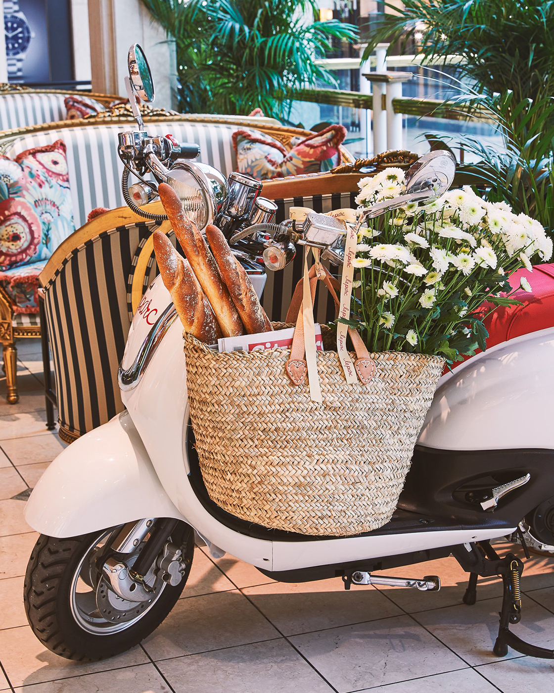 A white scooter with a woven basket attached to the front, filled with white flowers and baguettes, parked indoors near a seating area with patterned cushions and indoor plants.