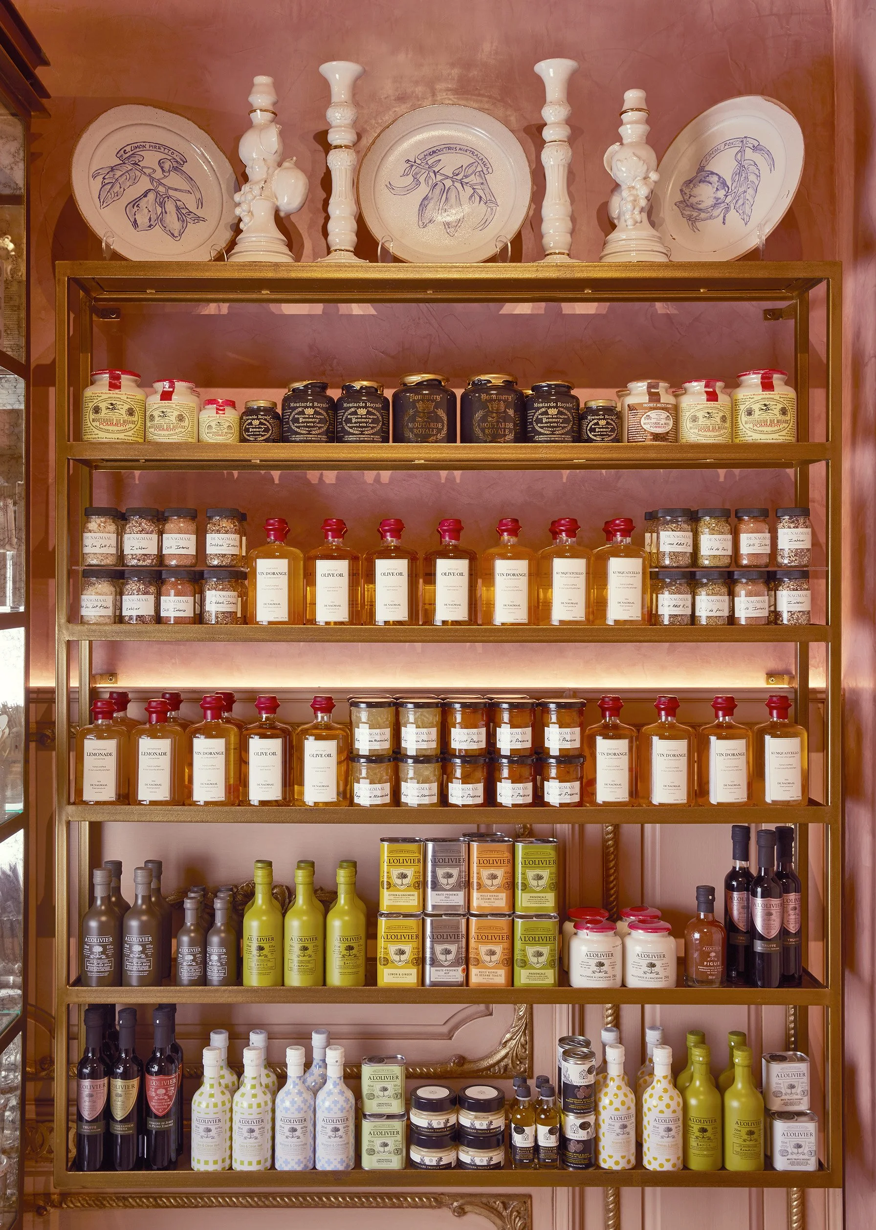 Shelving unit with various jars, bottles, and containers of gourmet food products, including olive oil, mustard, and condiments, against a pink wall with decorative plates and bust sculptures on the top shelf.