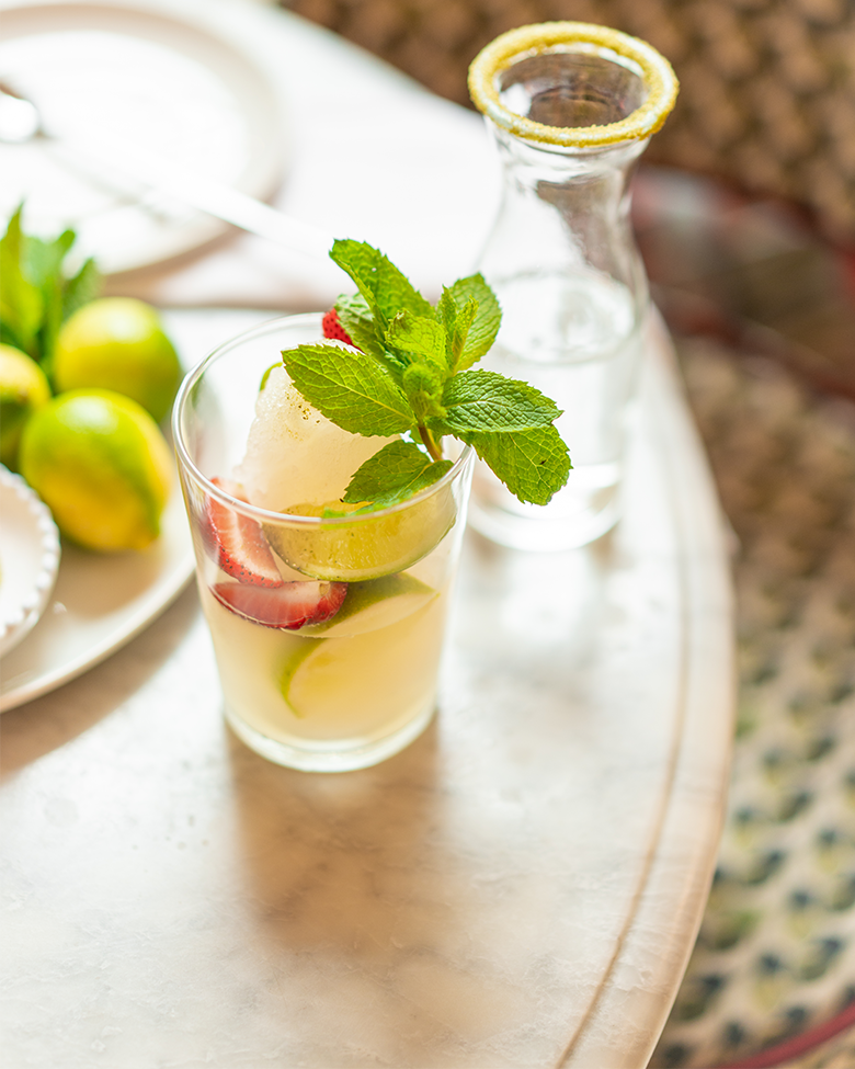 A glass of lemonade with slices of lime, strawberries, and mint leaves on top, next to a water carafe with a lemon wedge rim on a table.