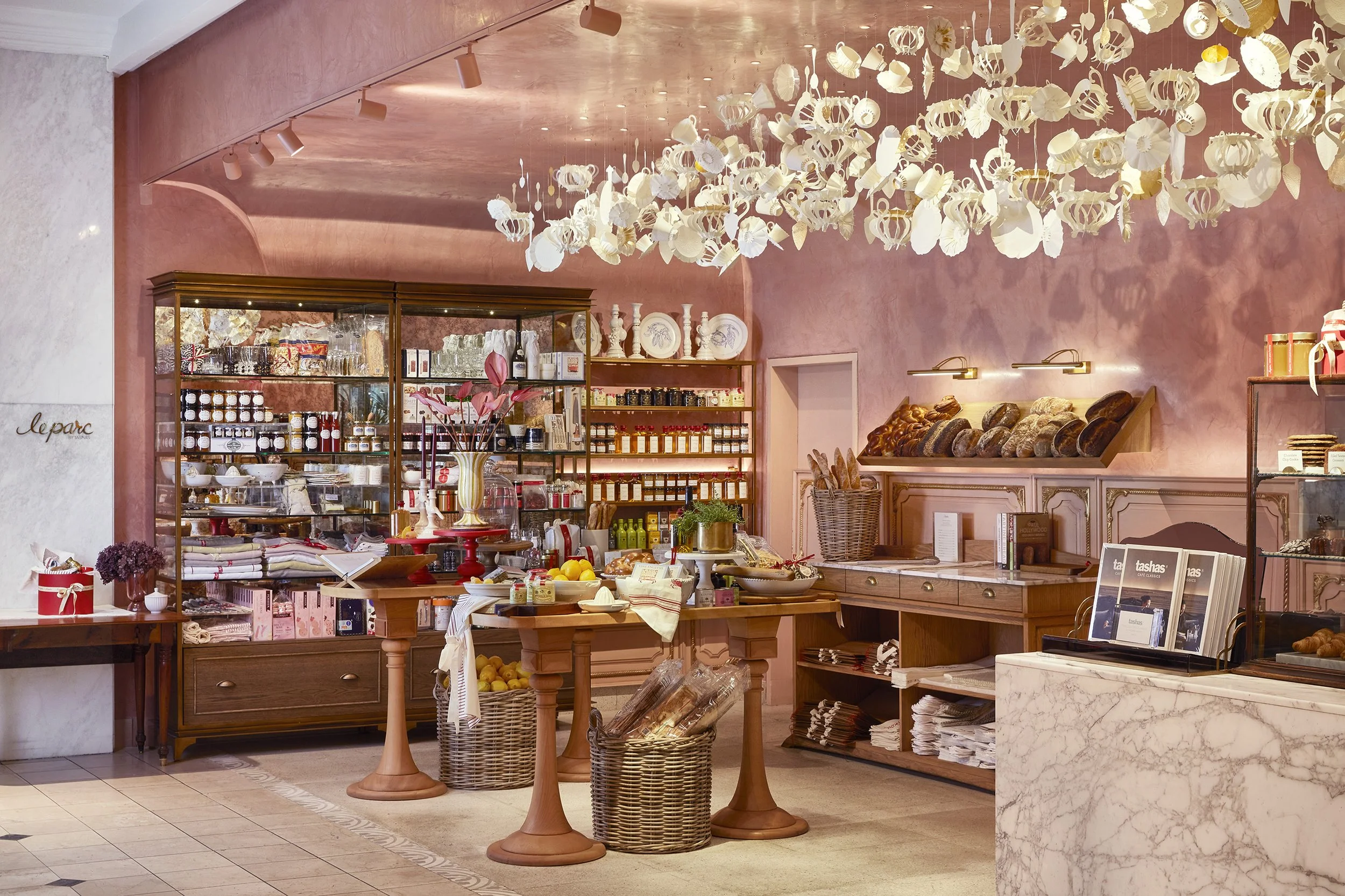 Interior of a bakery or cafe with pink walls, decorated with hanging paper art and filled with various baked goods and products on wooden shelves and tables.