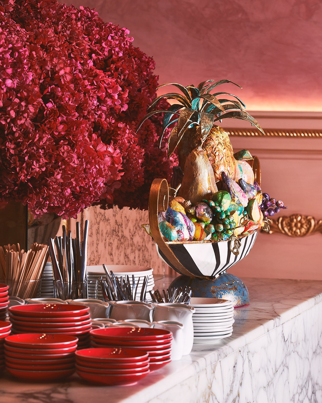 Colorful fruit sculpture in a bowl next to a large pink flower arrangement on a marble countertop.