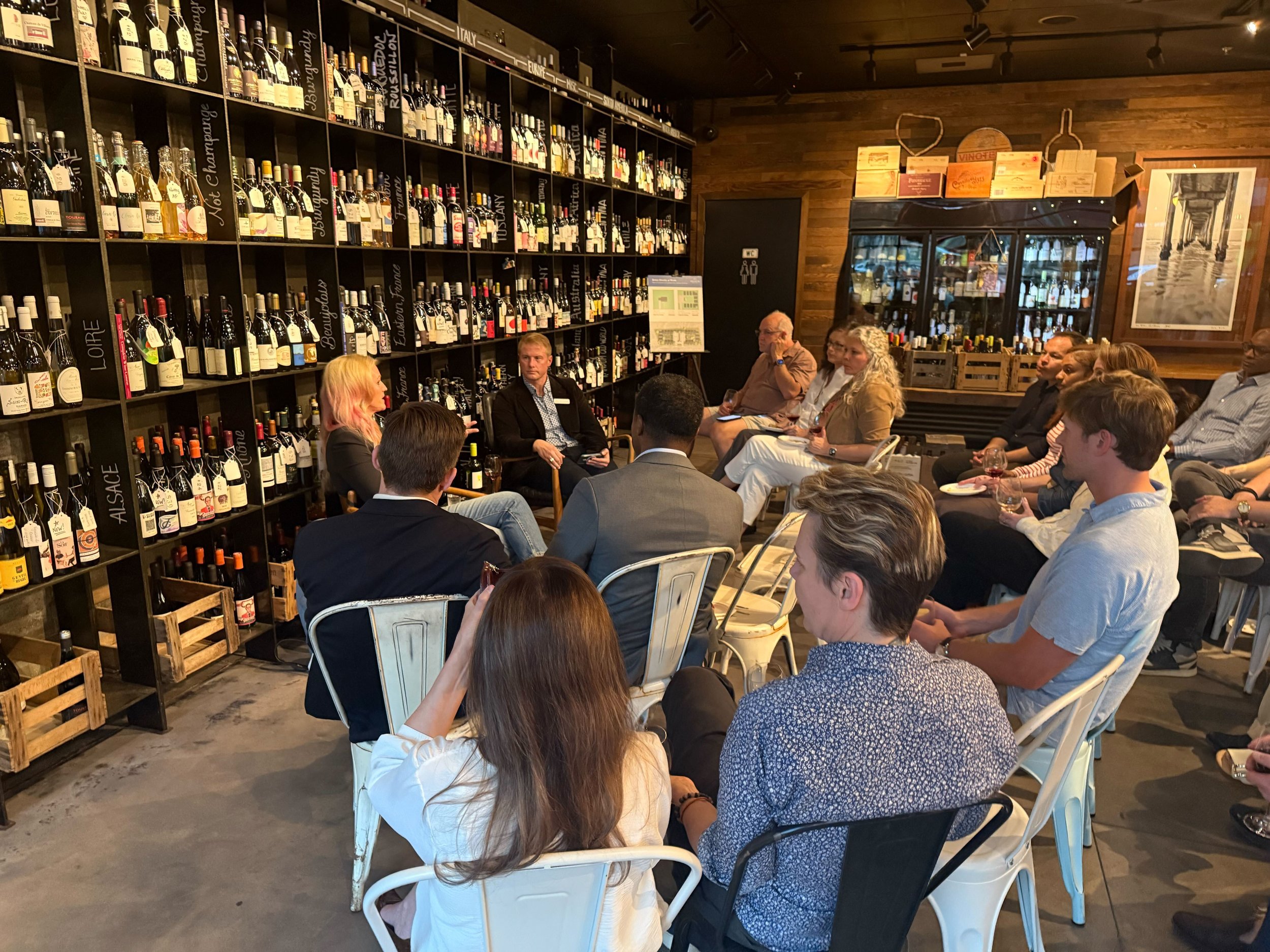 A group of people seated in a wine shop or tasting room, engaging in conversation. There are shelves filled with wine bottles on the left and a large mirror or window reflecting part of the room on the right. The atmosphere is cozy and casual, with wooden walls and dim lighting.