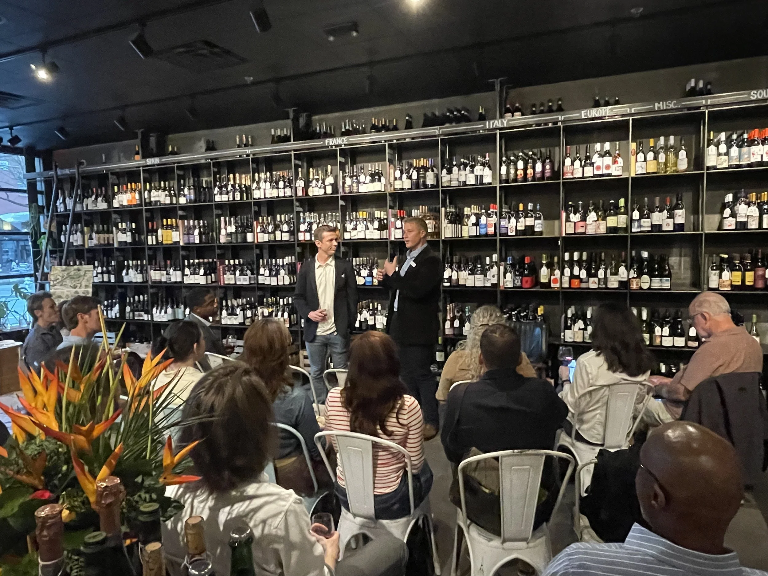 A group of people seated at tables in a restaurant or bar, with two men standing and talking in front of a wall of wine bottles categorized by country, including Spain, France, Italy, Europe, Misc., and South America.