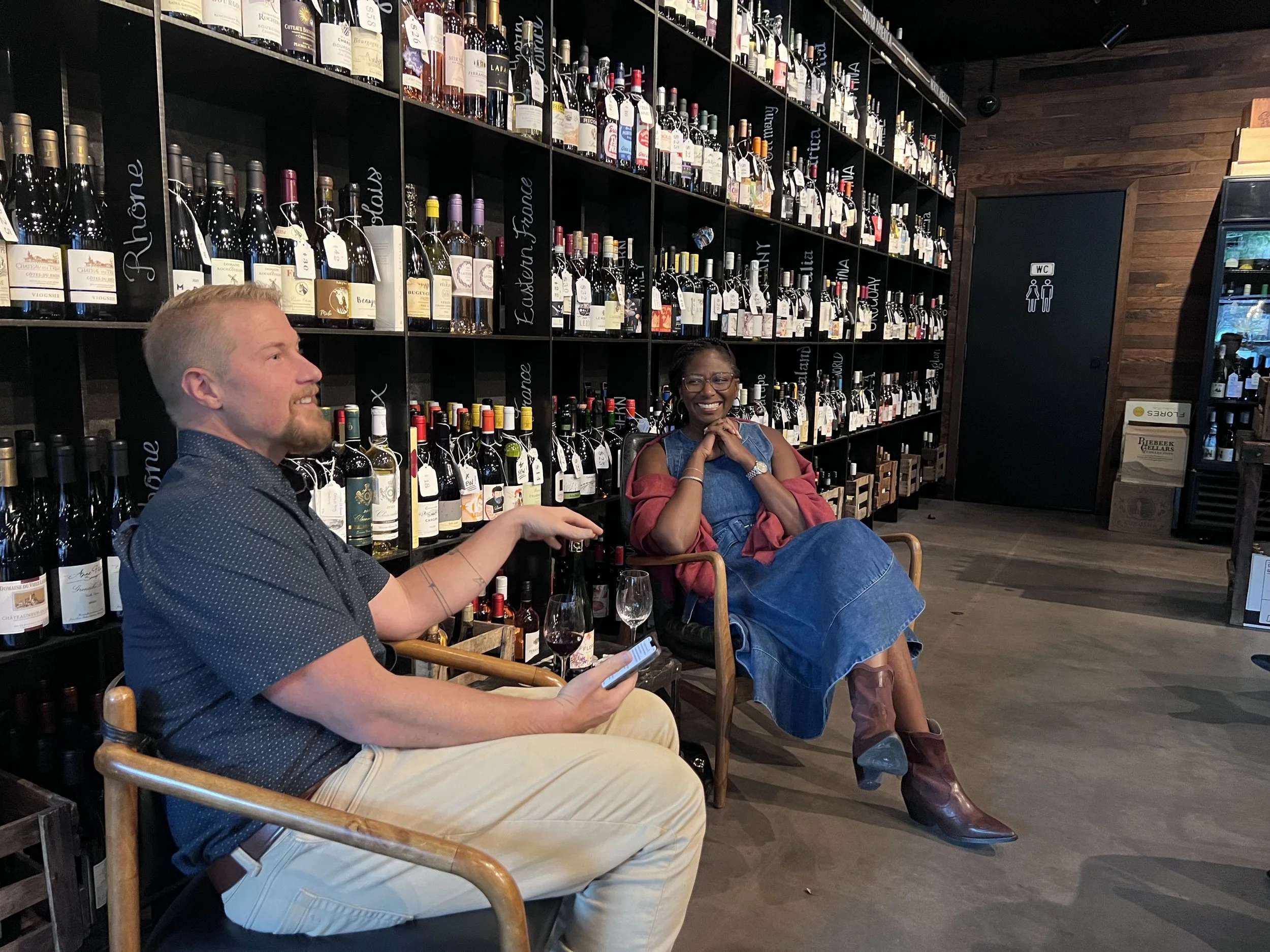 Two people sitting at a table in a wine store or wine bar, with shelves filled with wine bottles behind them. The man on the left, with light-colored hair and a beard, is smiling and looking to his right while using his phone. The woman on the right, with dark hair and glasses, is smiling, sitting with her hands clasped under her chin. She is wearing a denim shirt and brown boots, with her legs crossed.
